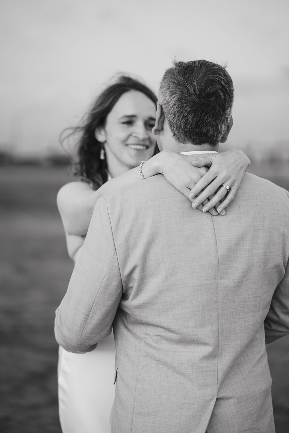 Black and white photo of a smiling bride embracing a groom from behind outdoors. Oceanside, CA