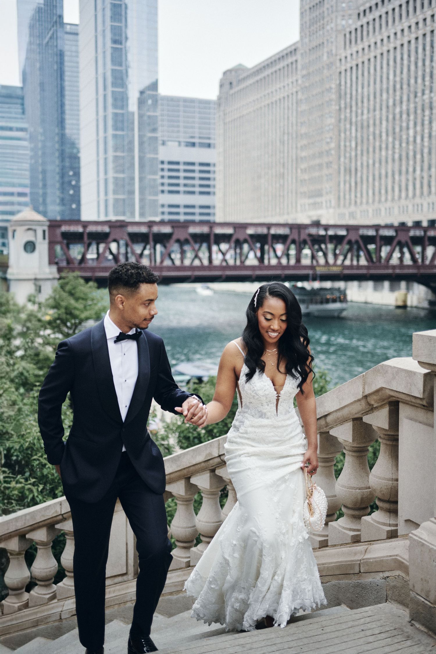 Couple in wedding attire walks down stone steps by Chicago River with city skyline and bridge backdrop.