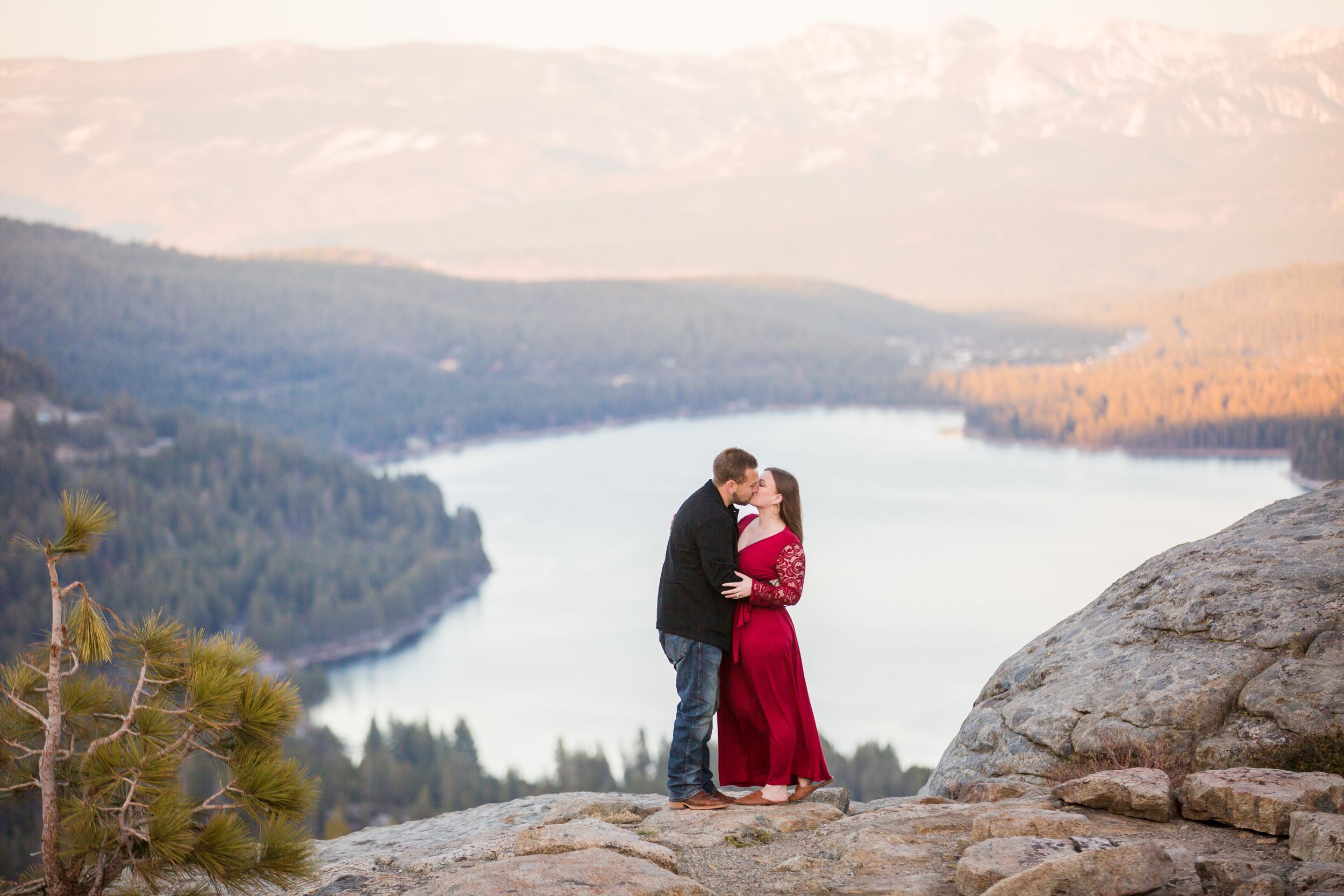 Couple sharing a romantic kiss on a rocky mountain overlook with a stunning lake and forest backdrop at sunset.