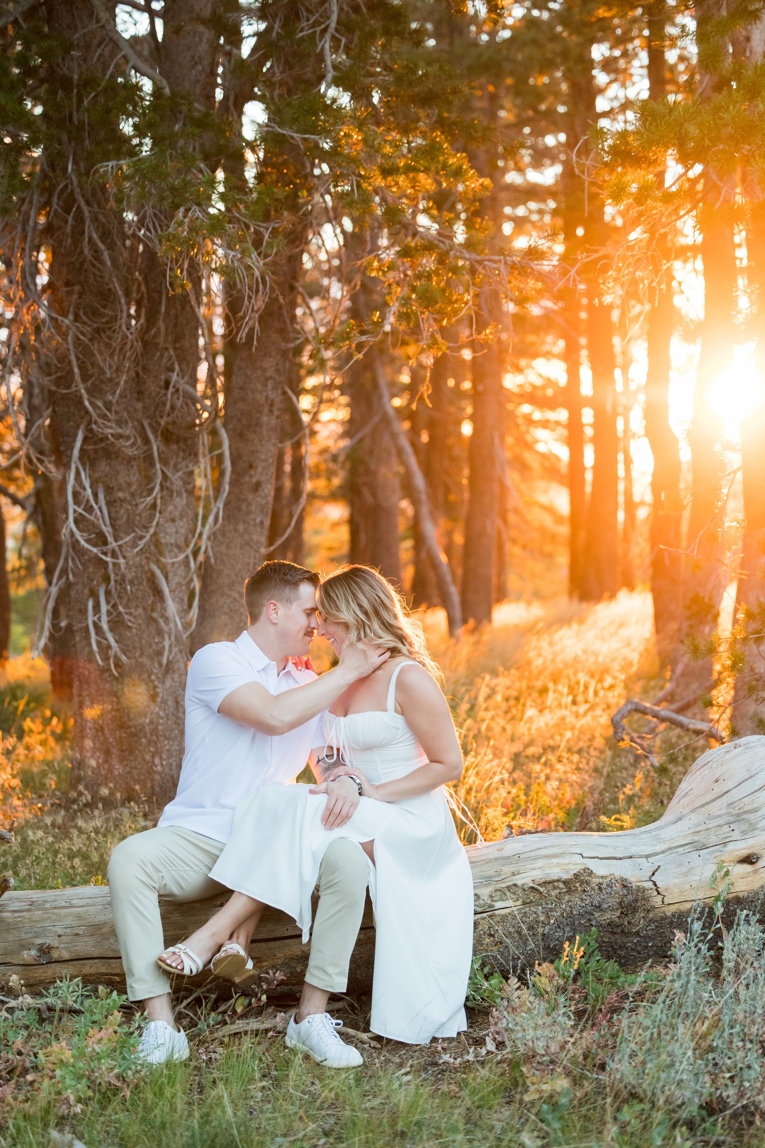 Couple sitting on a log in a sunlit forest at golden hour, sharing a romantic moment in white outfits.