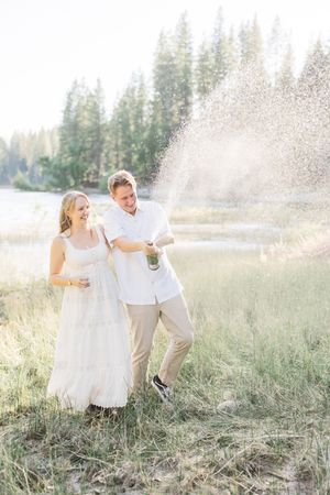 Couple in white celebrating outdoors by a lake, popping champagne in a meadow surrounded by tall pine trees.