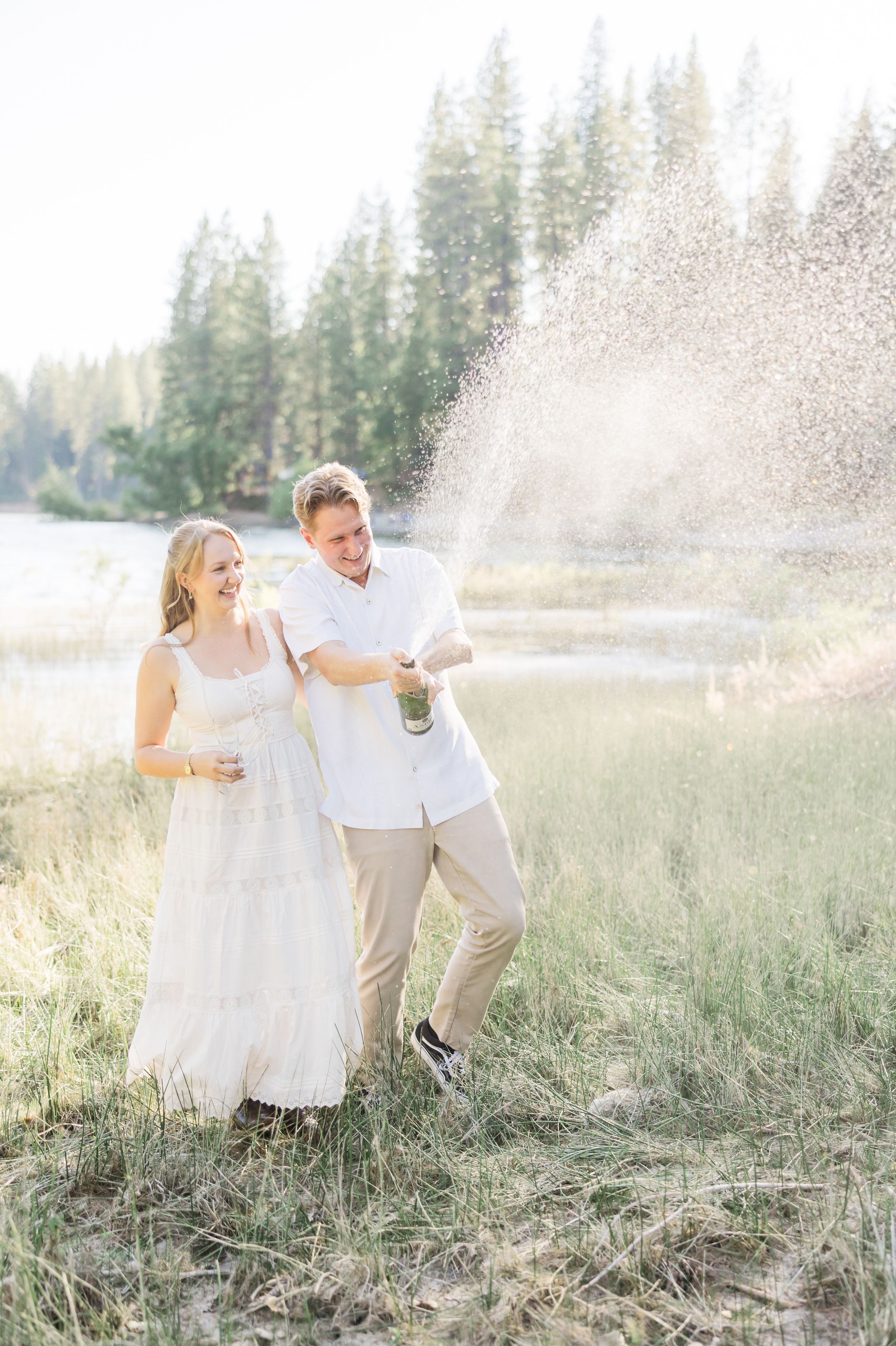 Couple in white celebrating outdoors by a lake, popping champagne in a meadow surrounded by tall pine trees.