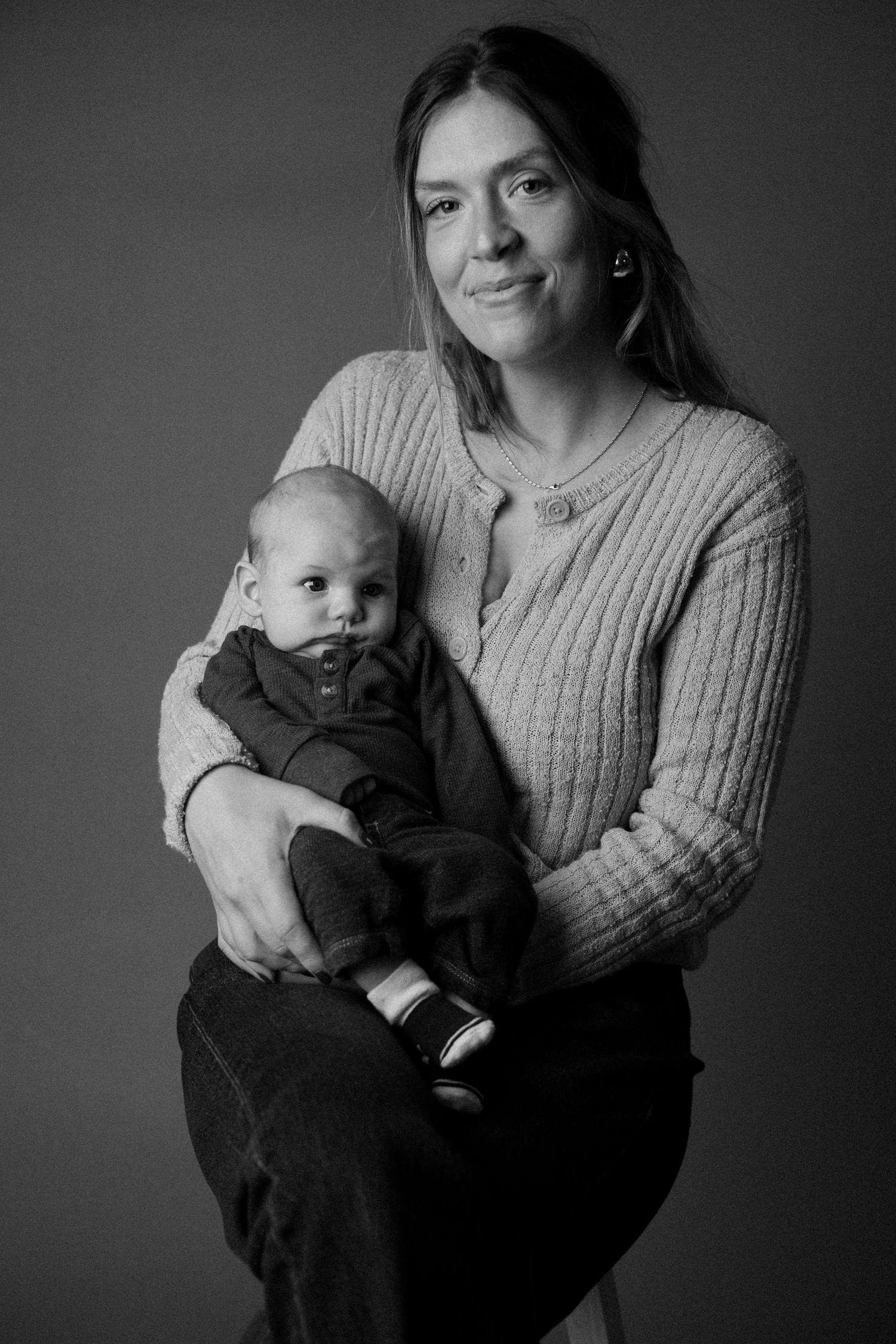 Black and white portrait of a smiling woman holding a baby in a knit sweater against a gray background.