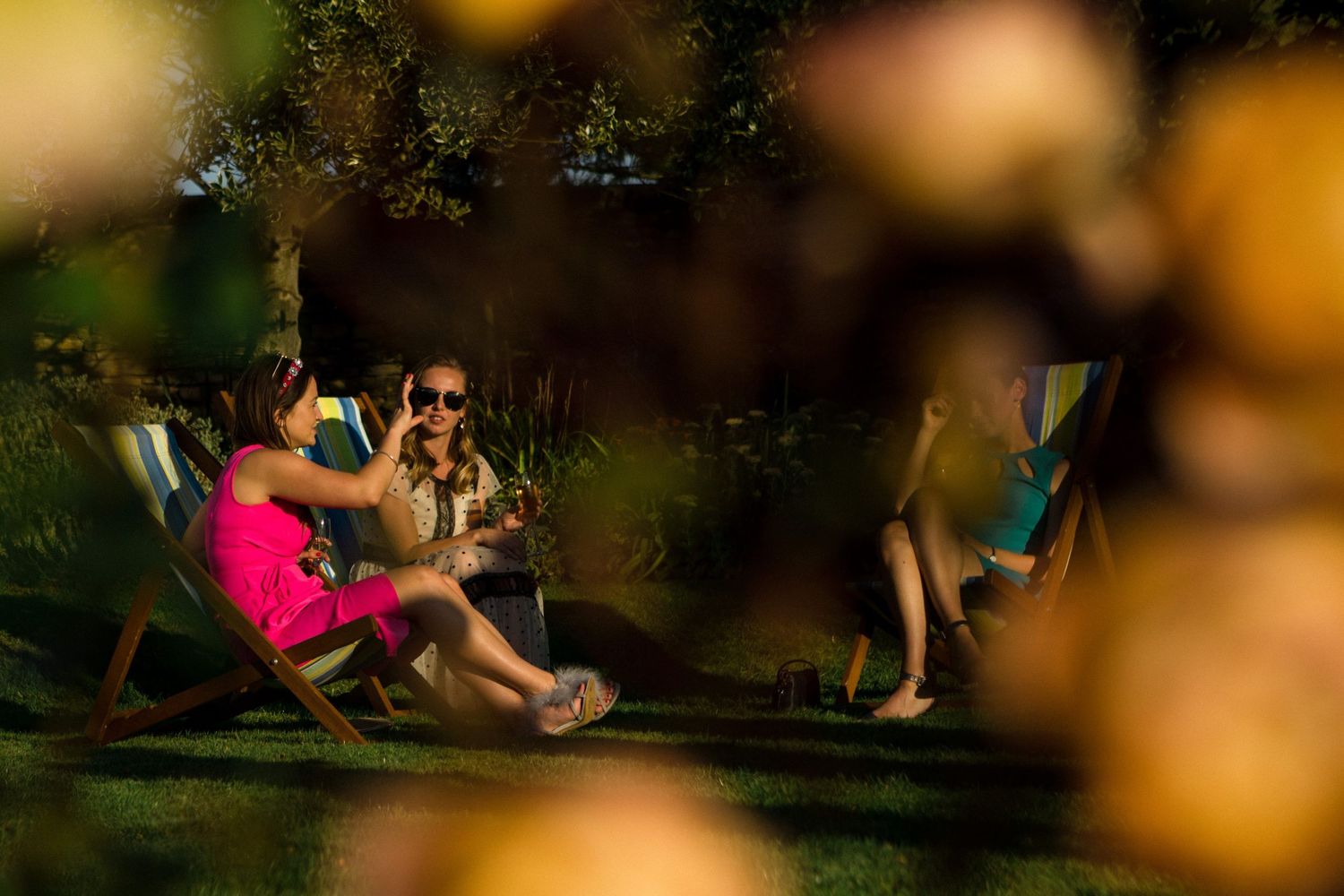 Two women in summer dresses enjoy drinks in garden chairs outdoors, framed by bokeh foliage.