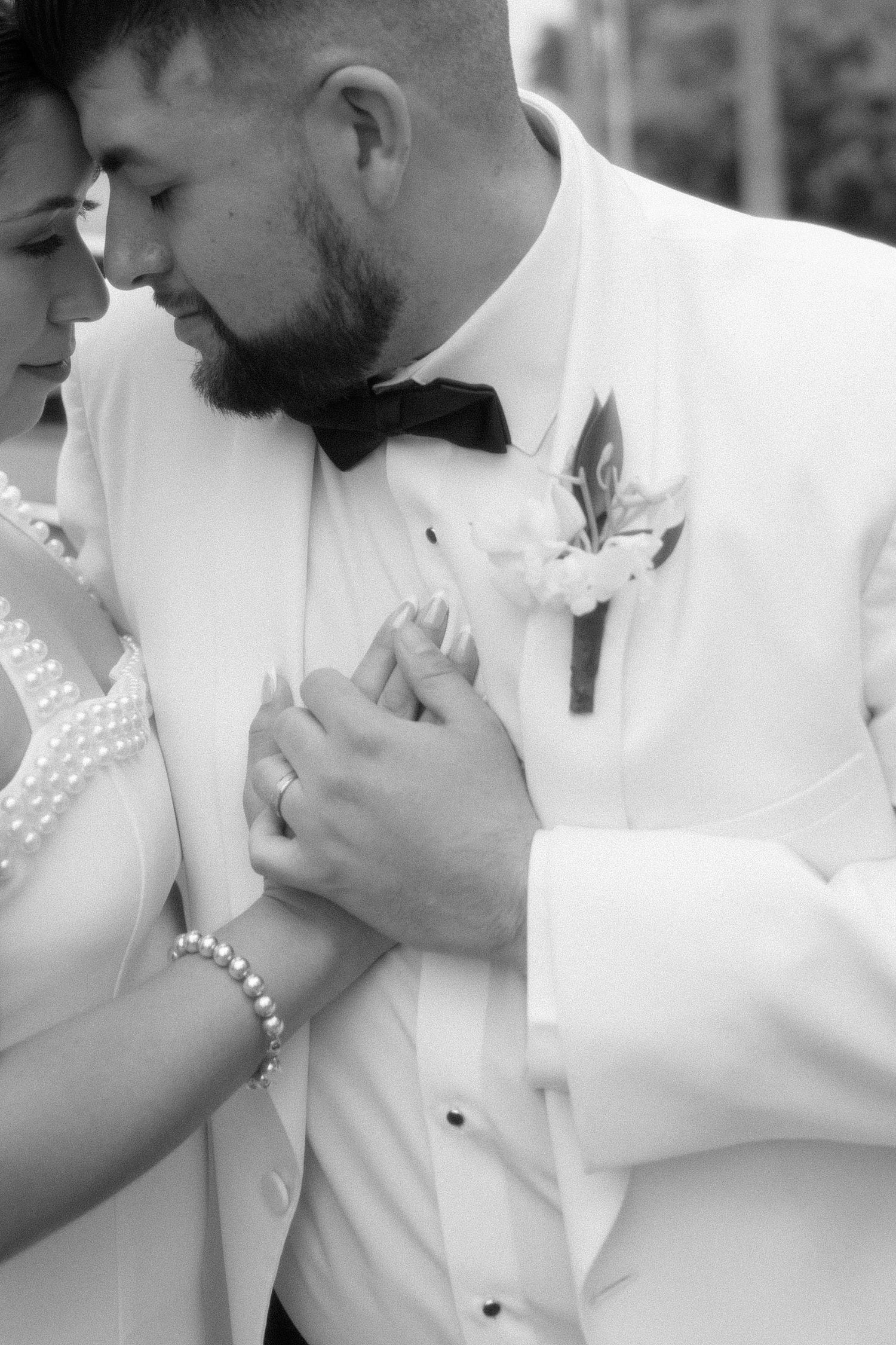 Black and white wedding photo of bride and groom holding hands, groom in white tuxedo with boutonniere and bow tie.