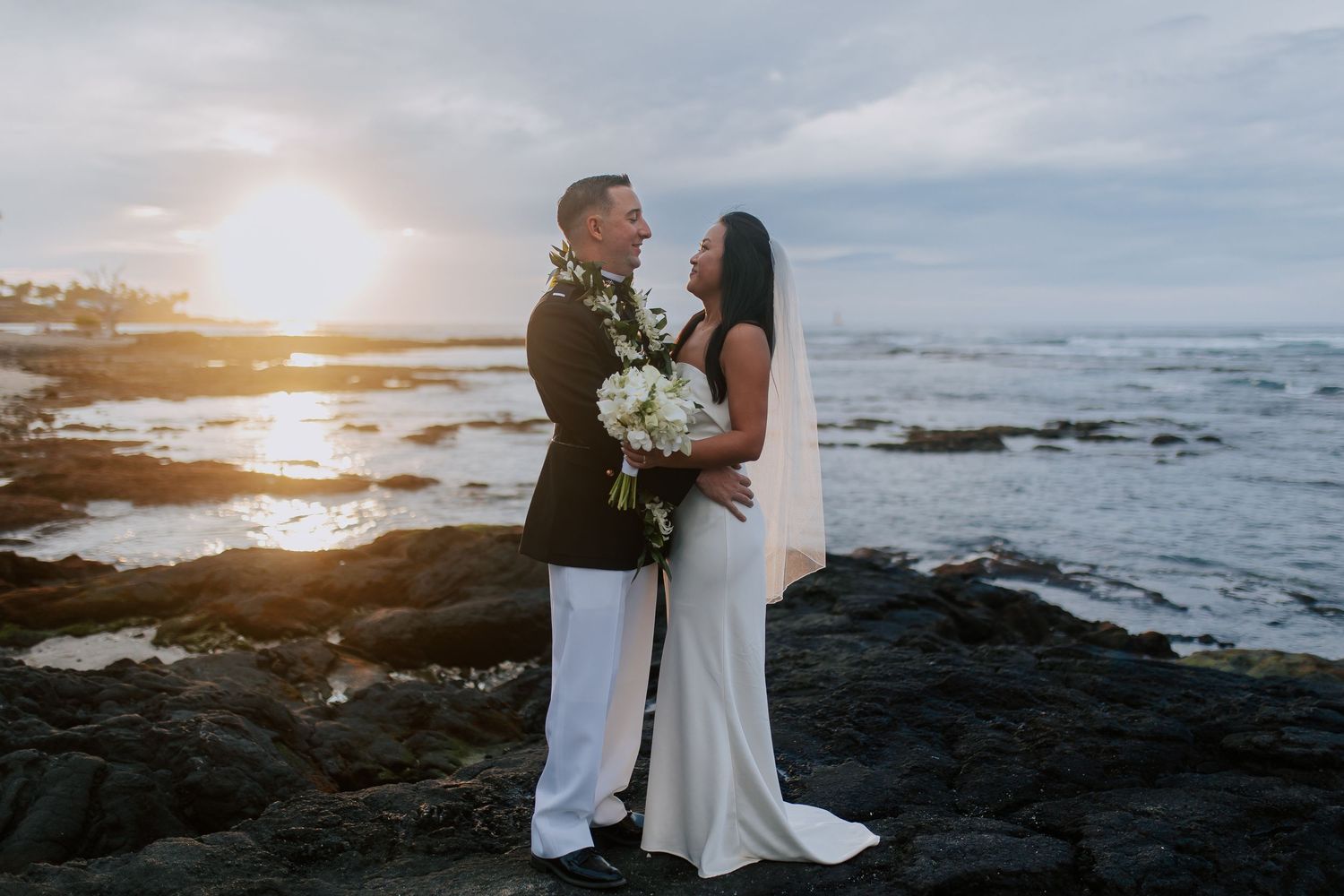 Couple embracing at sunset on rocky Hawaiian coast, bride in white gown holding bouquet, groom in military dress uniform.