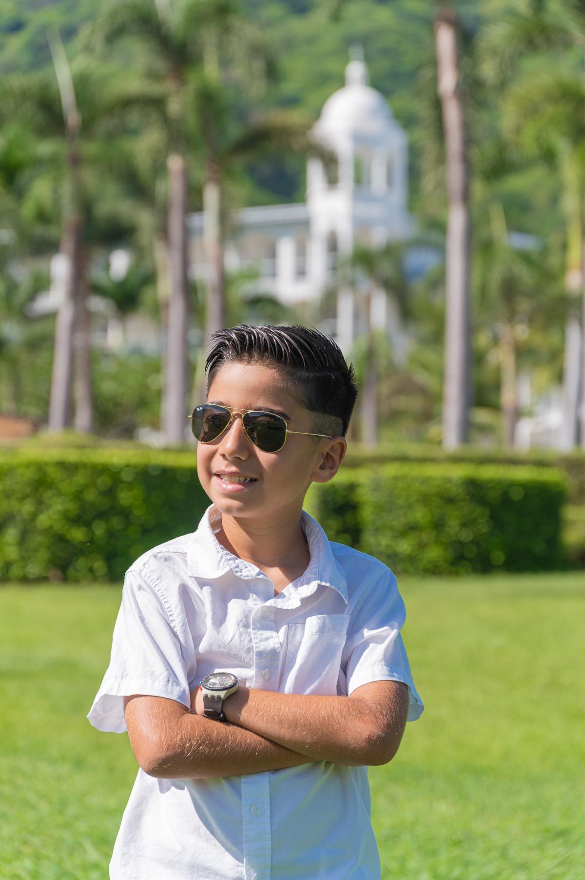 Smiling boy in white shirt and sunglasses stands with arms crossed in a lush green park with palm trees at Riu Palace, Guanacaste, Costa Ric