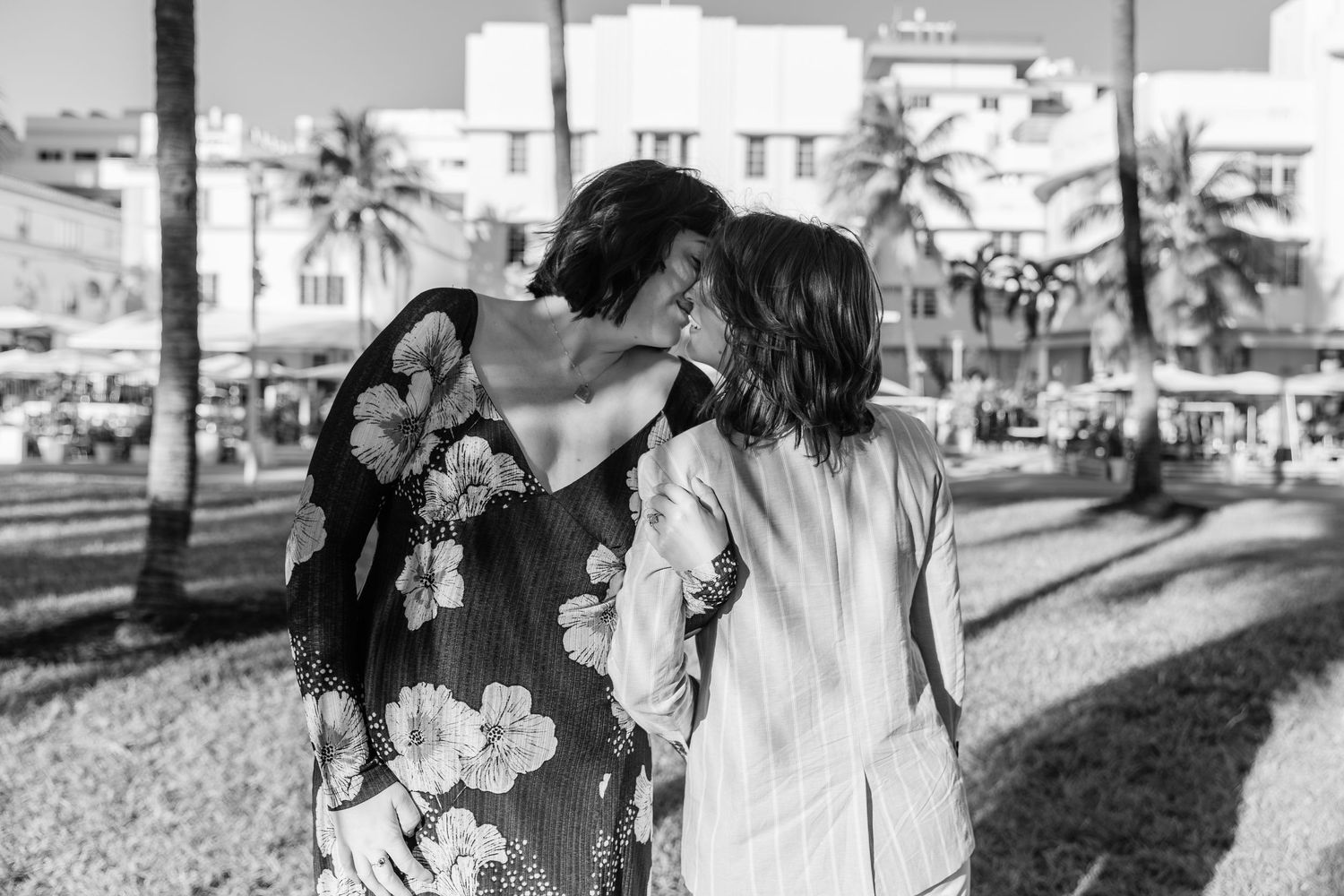 Black and white photo of two women sharing a kiss outdoors near palm trees and buildings in a sunny plaza.