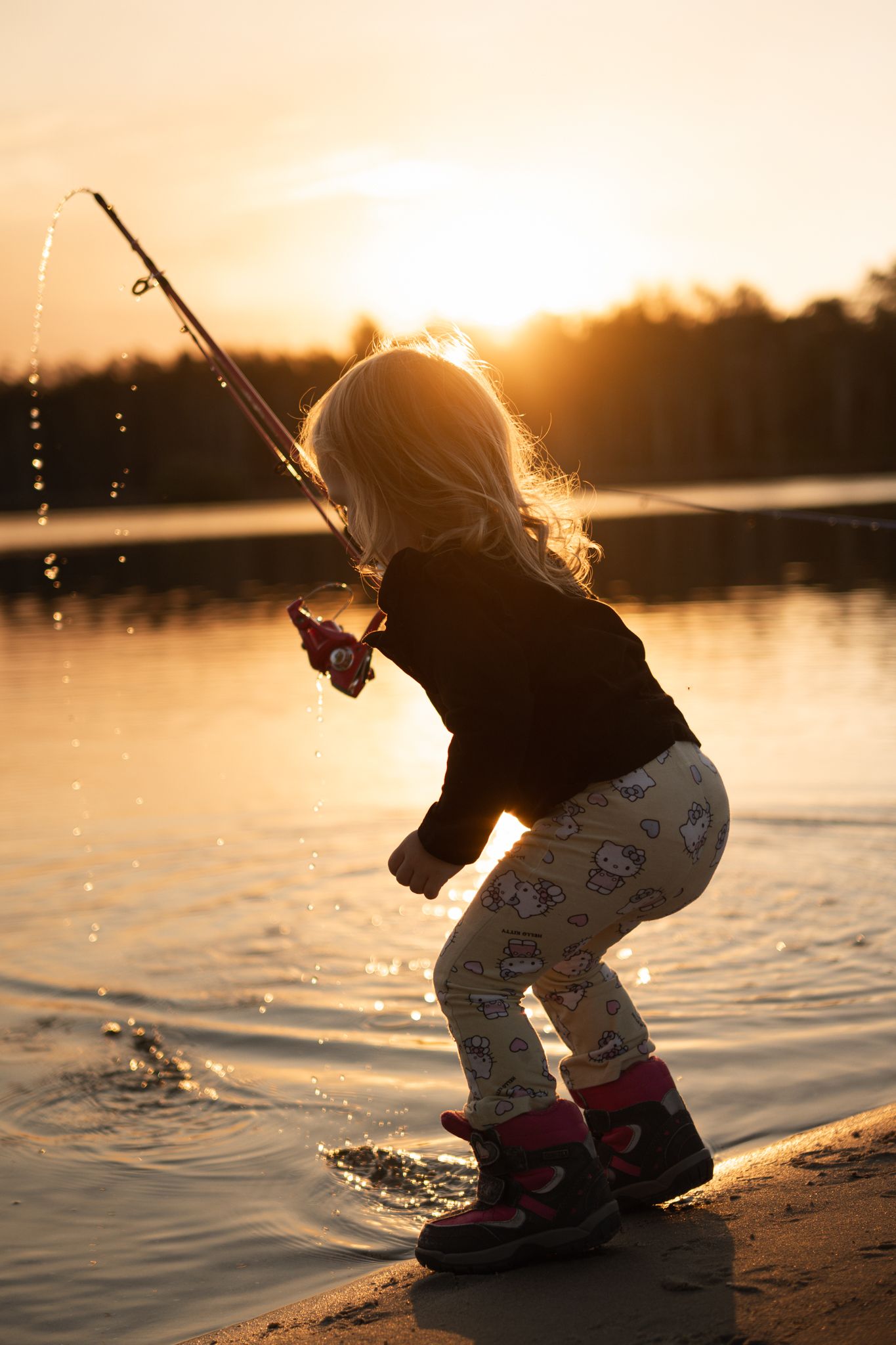 Young blonde girl fishing at golden sunset by a lake, pulling her rod back as water splashes around her boots.