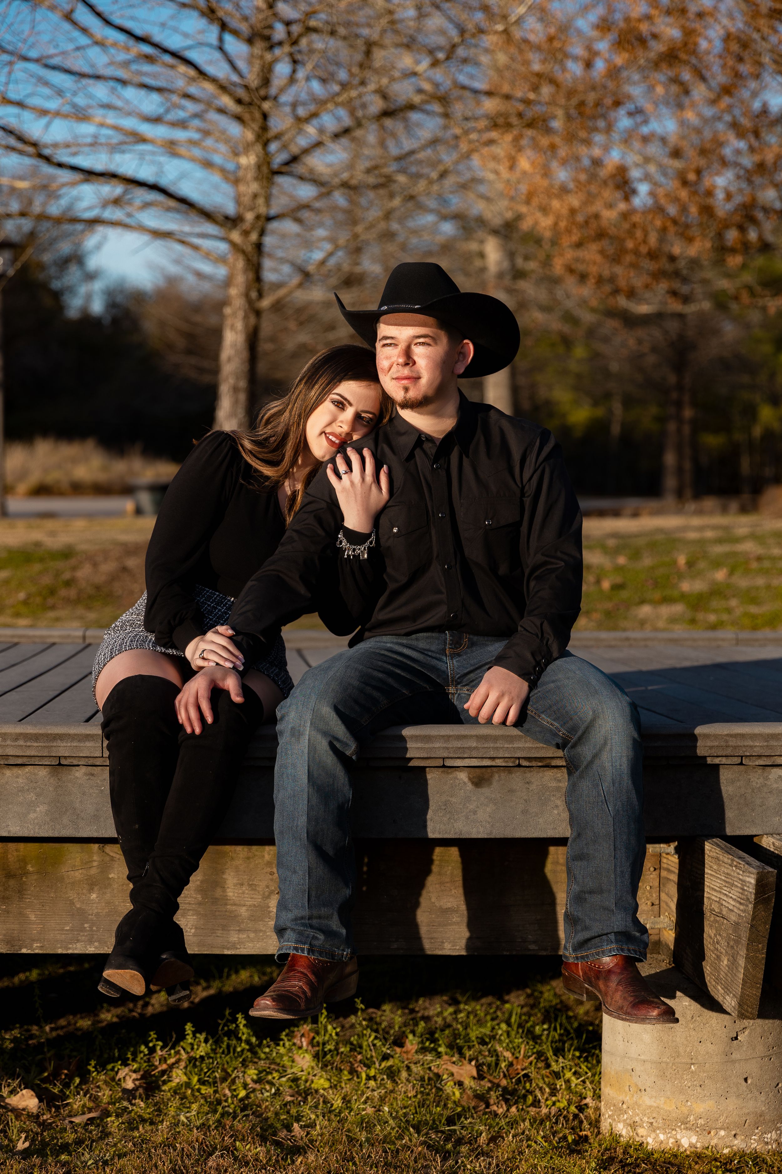 A couple in black outfits sits on a wooden bench outdoors, man wearing cowboy hat, woman leaning on his shoulder.