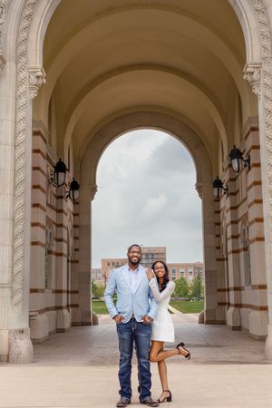 A couple poses under an ornate stone archway, the man in a blue blazer and jeans, the woman in a white dress.