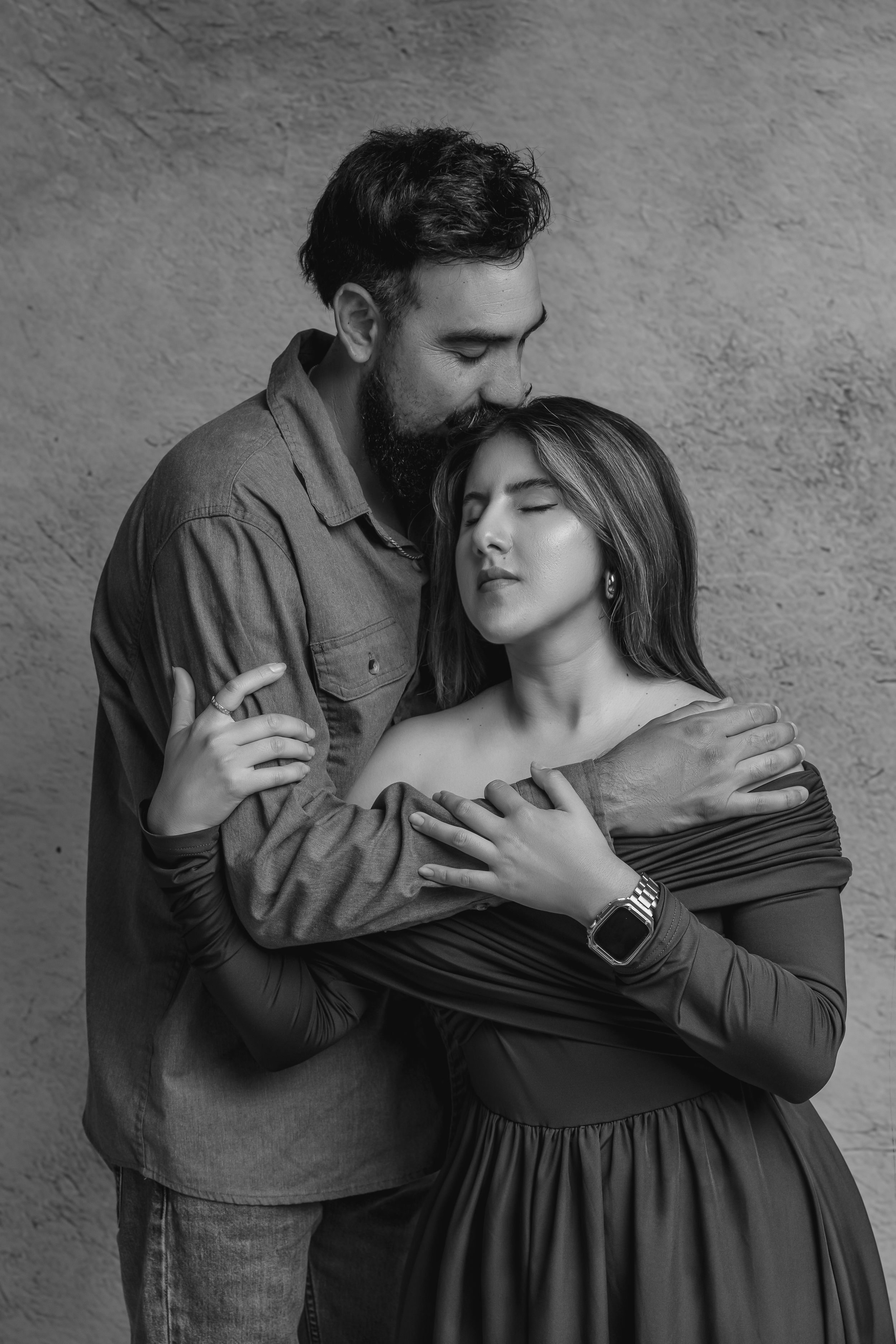 Black and white photo of a bearded man tenderly embracing a woman from behind, kissing her head in a romantic pose.