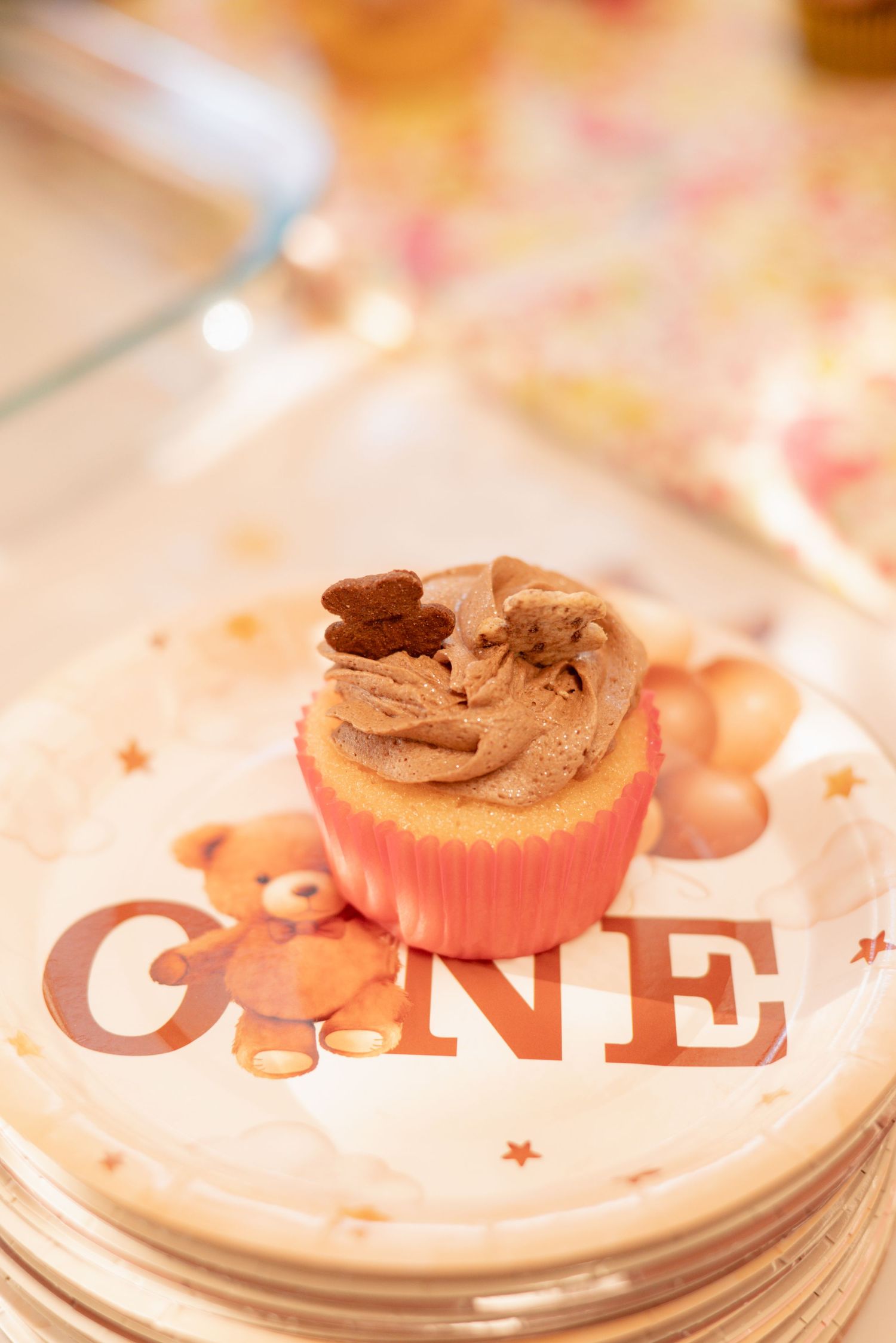 A chocolate frosted cupcake in a pink liner on a teddy bear first birthday plate reading ONE.