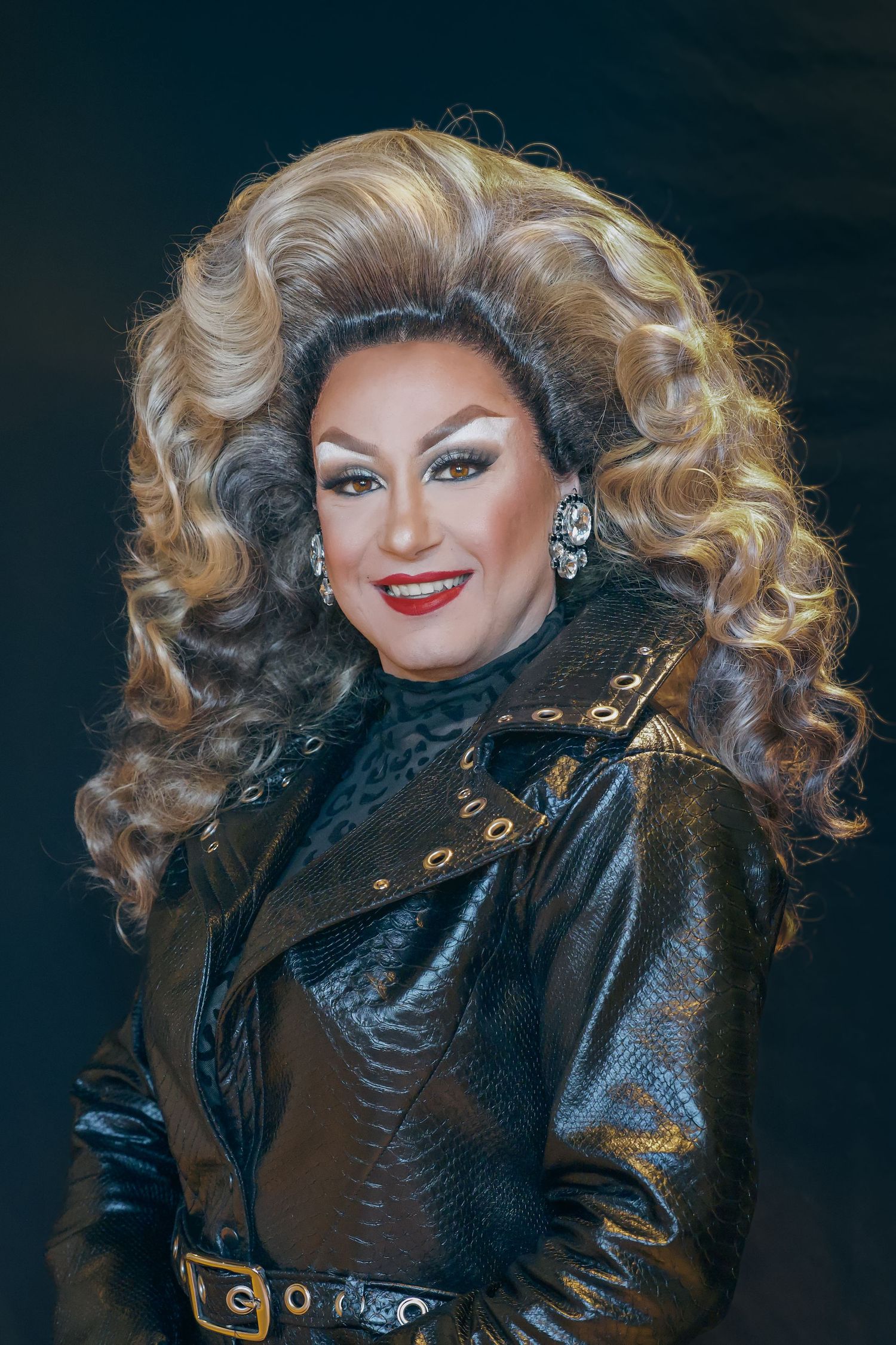 Drag performer in voluminous blonde wig, dramatic makeup, and black leather jacket poses confidently against dark background.