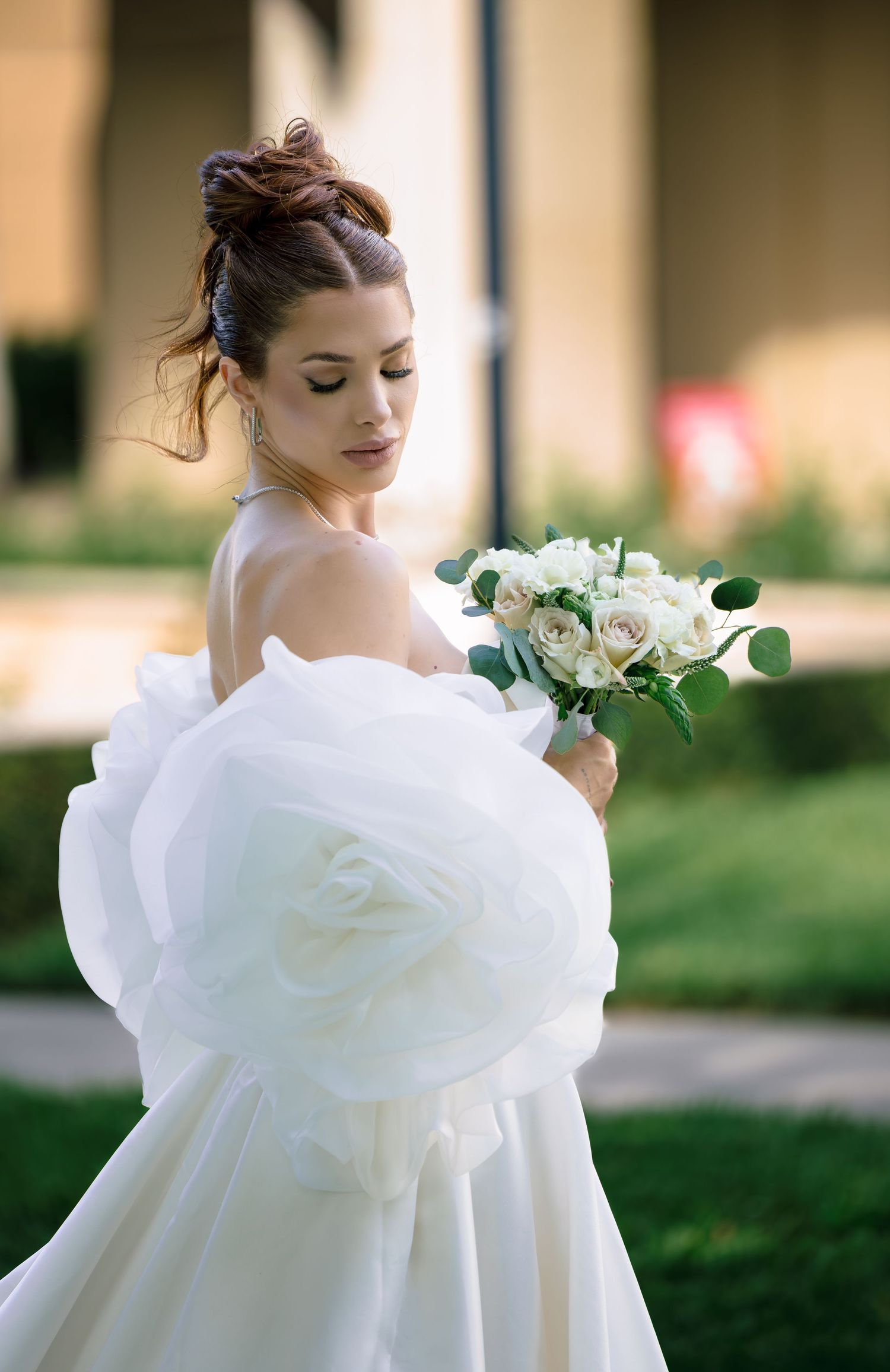 Bride in off-shoulder white ballgown with large floral detail holds white rose bouquet outdoors.