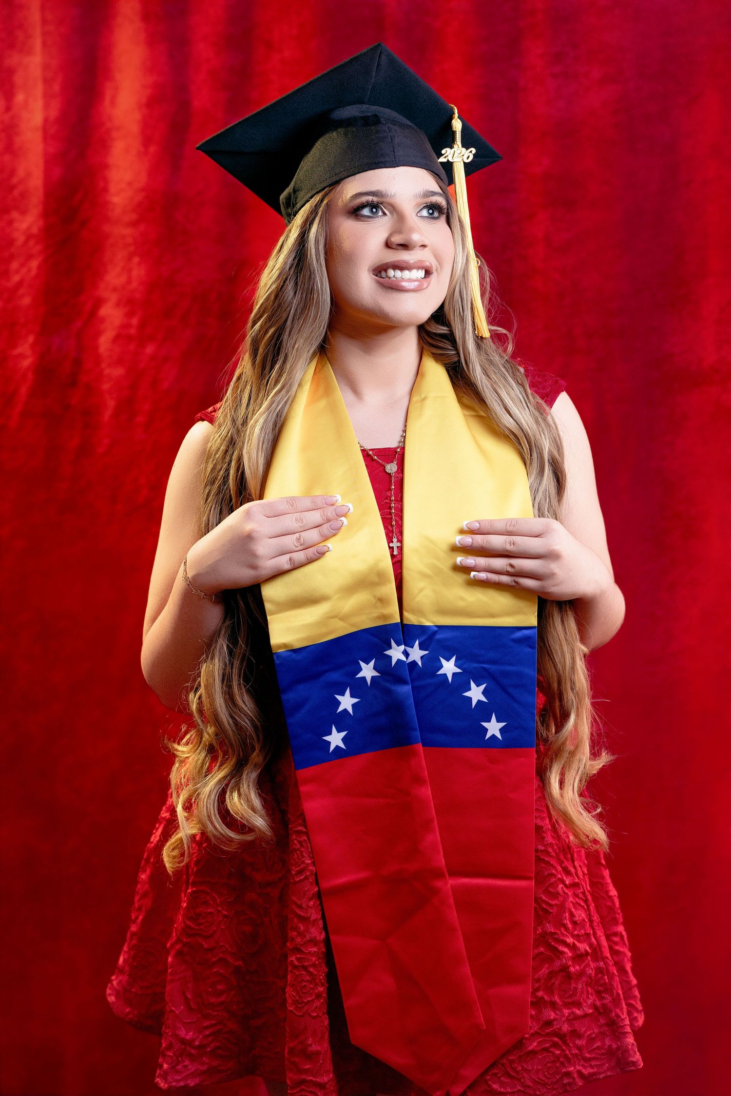 Young woman in red lace dress and black graduation cap and gown posing against red background.