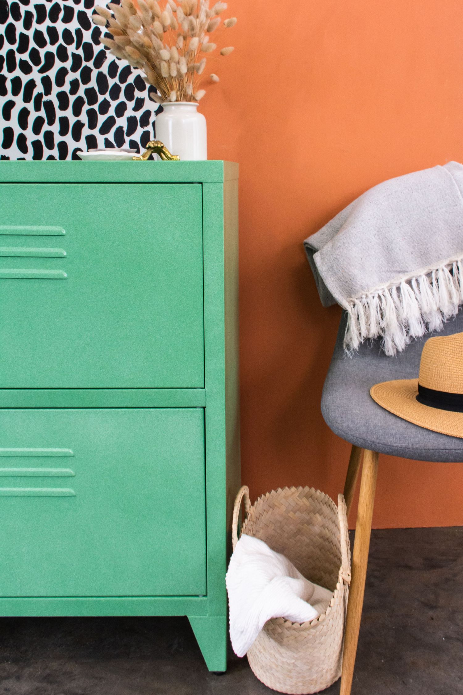 Green metal locker dresser beside orange wall with woven basket, gray chair, straw hat, and decorative vase.