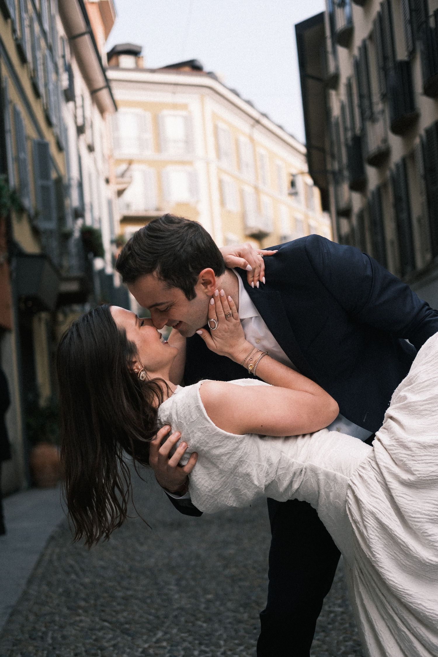 A couple shares a romantic dip kiss on a charming European cobblestone street, the groom in navy suit, bride in white dress.