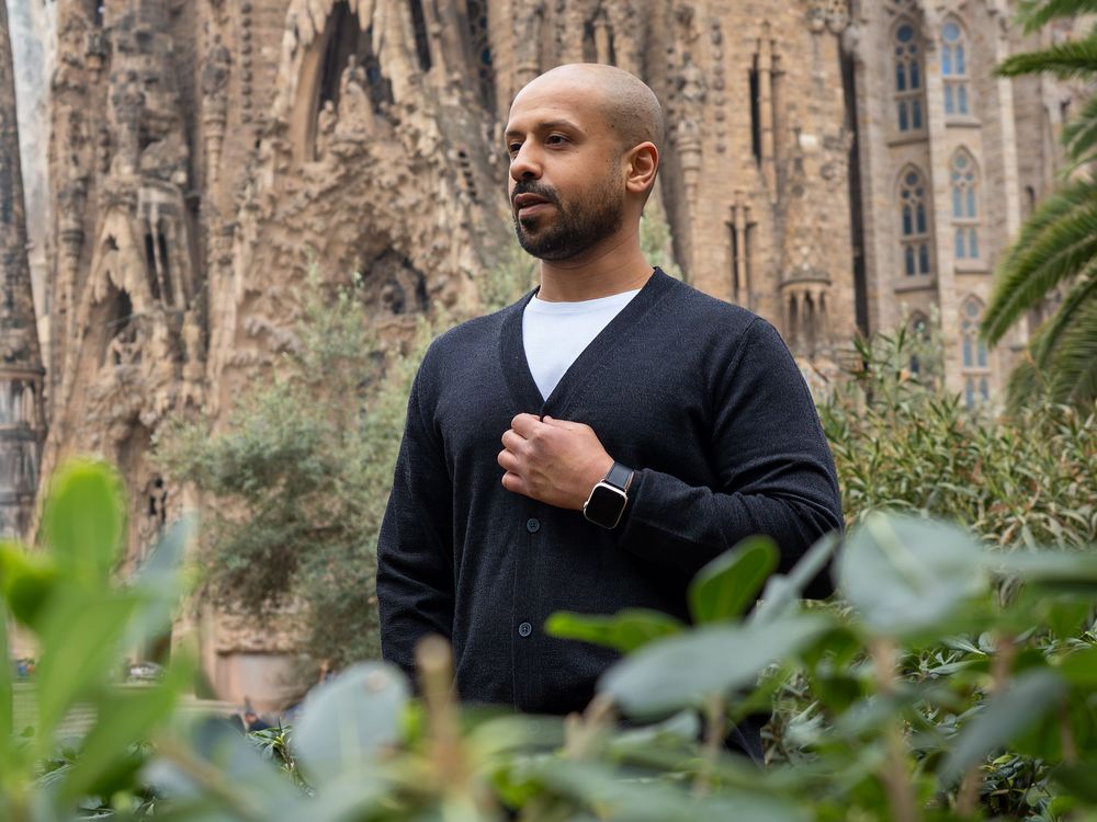 Man in black cardigan stands among greenery with Sagrada Familia cathedral in Barcelona visible behind him.