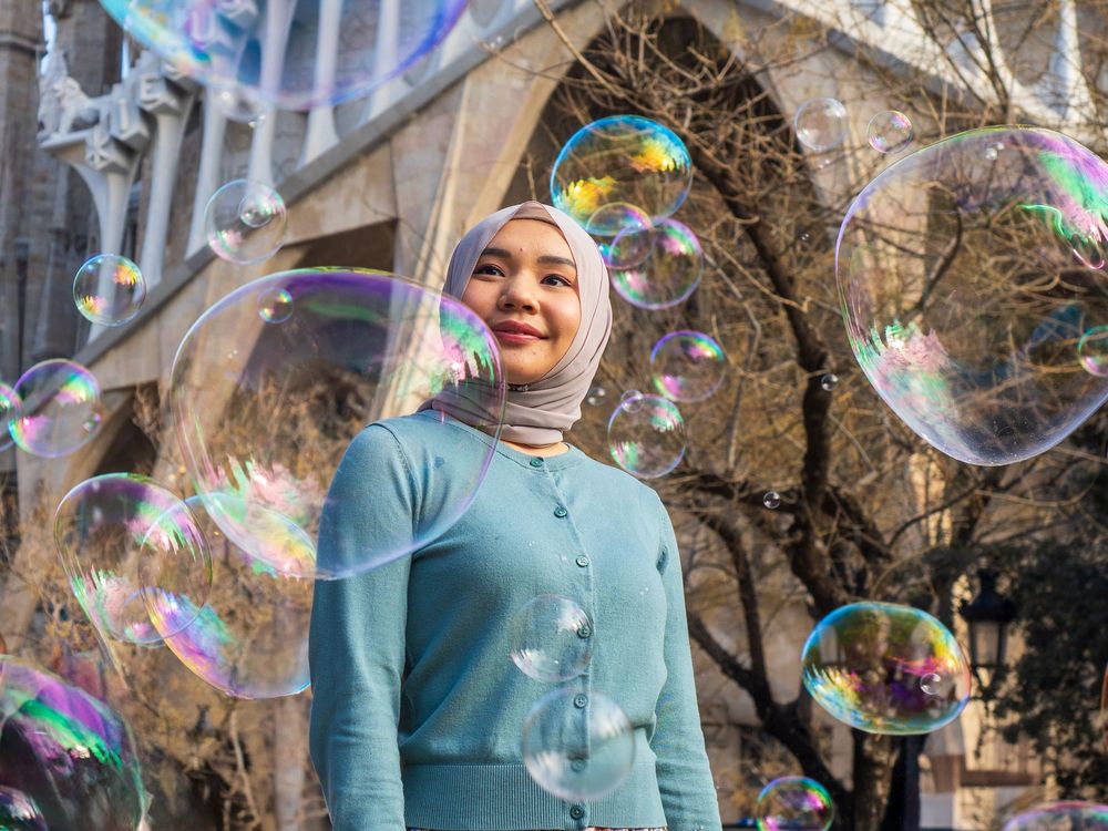 A young woman in a blue hijab and cardigan smiles surrounded by colorful soap bubbles outdoors.