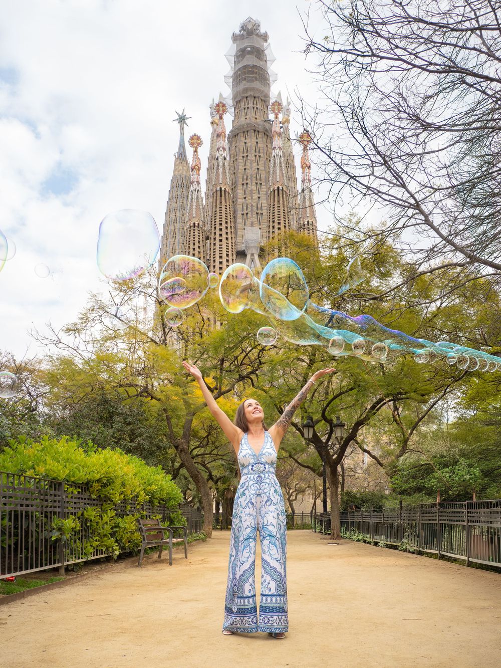 Person in blue and white patterned dress poses joyfully near the iconic Sagrada Familia cathedral in Barcelona, Spain.