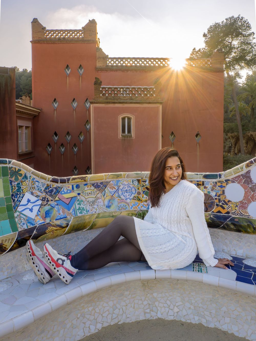 A mosaic-tiled bench curves along a terracotta wall with sunlight creating a warm glow at Park Guell in Barcelona.
