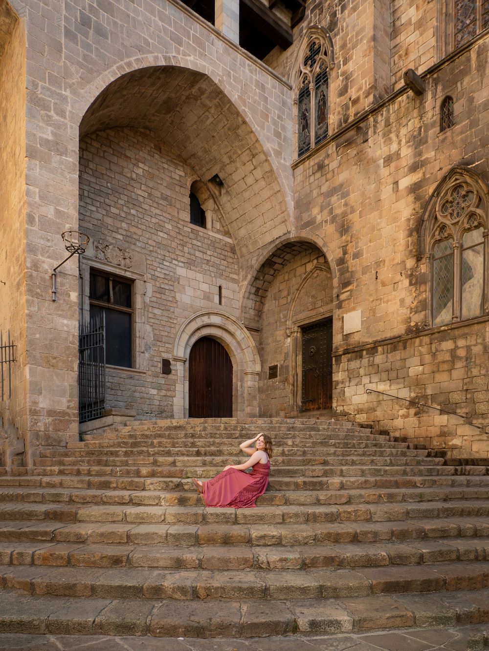 Woman in red dress poses gracefully on ancient stone steps beside a Gothic arch in Barcelona's historic quarter.