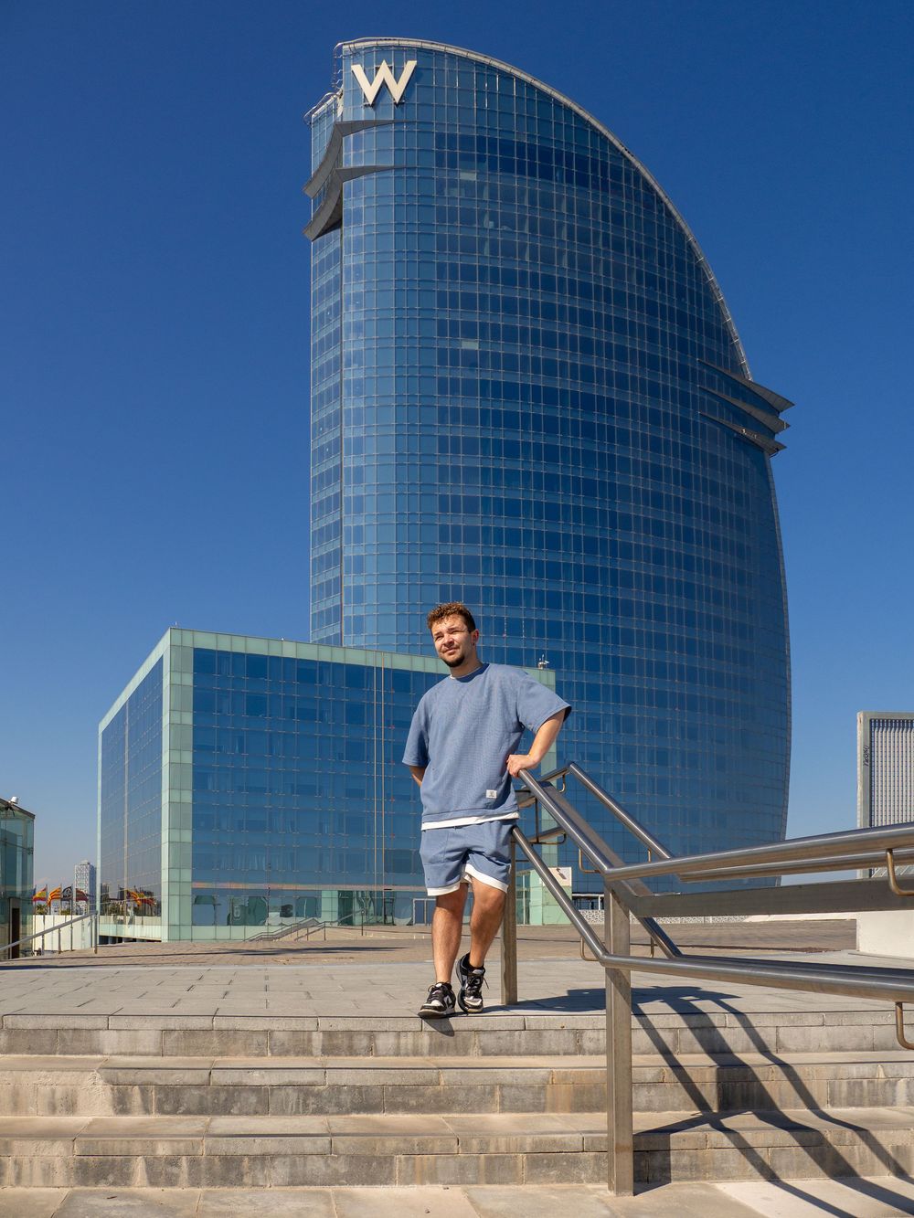 Young man posing on steps in front of the iconic W Hotel skyscraper in Barcelona on a sunny day.
