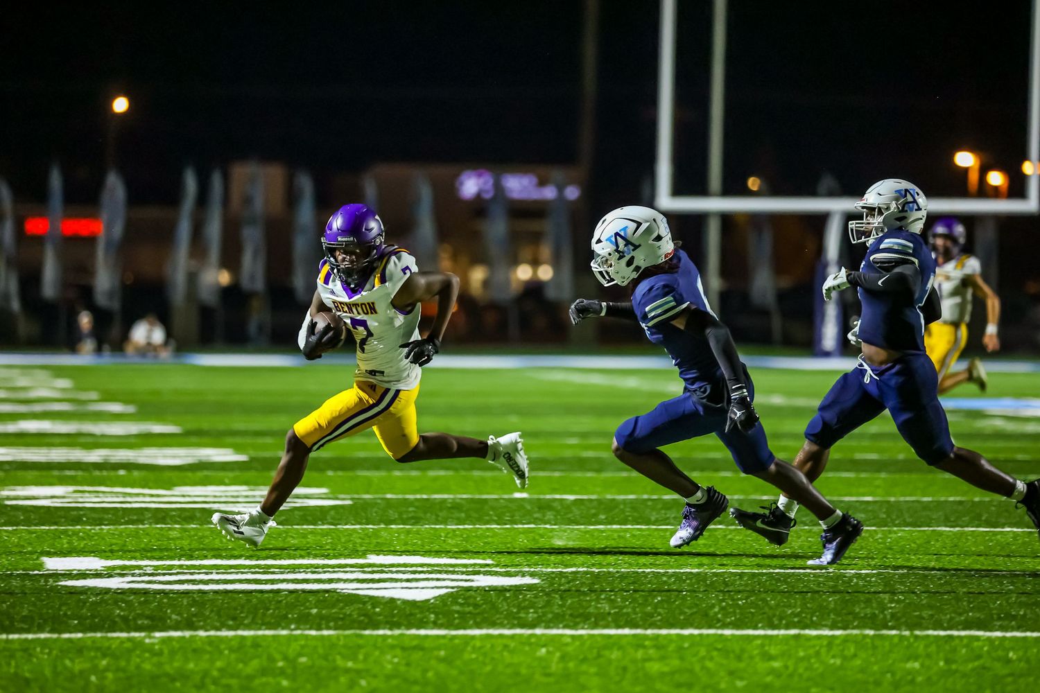 High school football player in yellow jersey runs with the ball while being chased by defenders in blue uniforms.