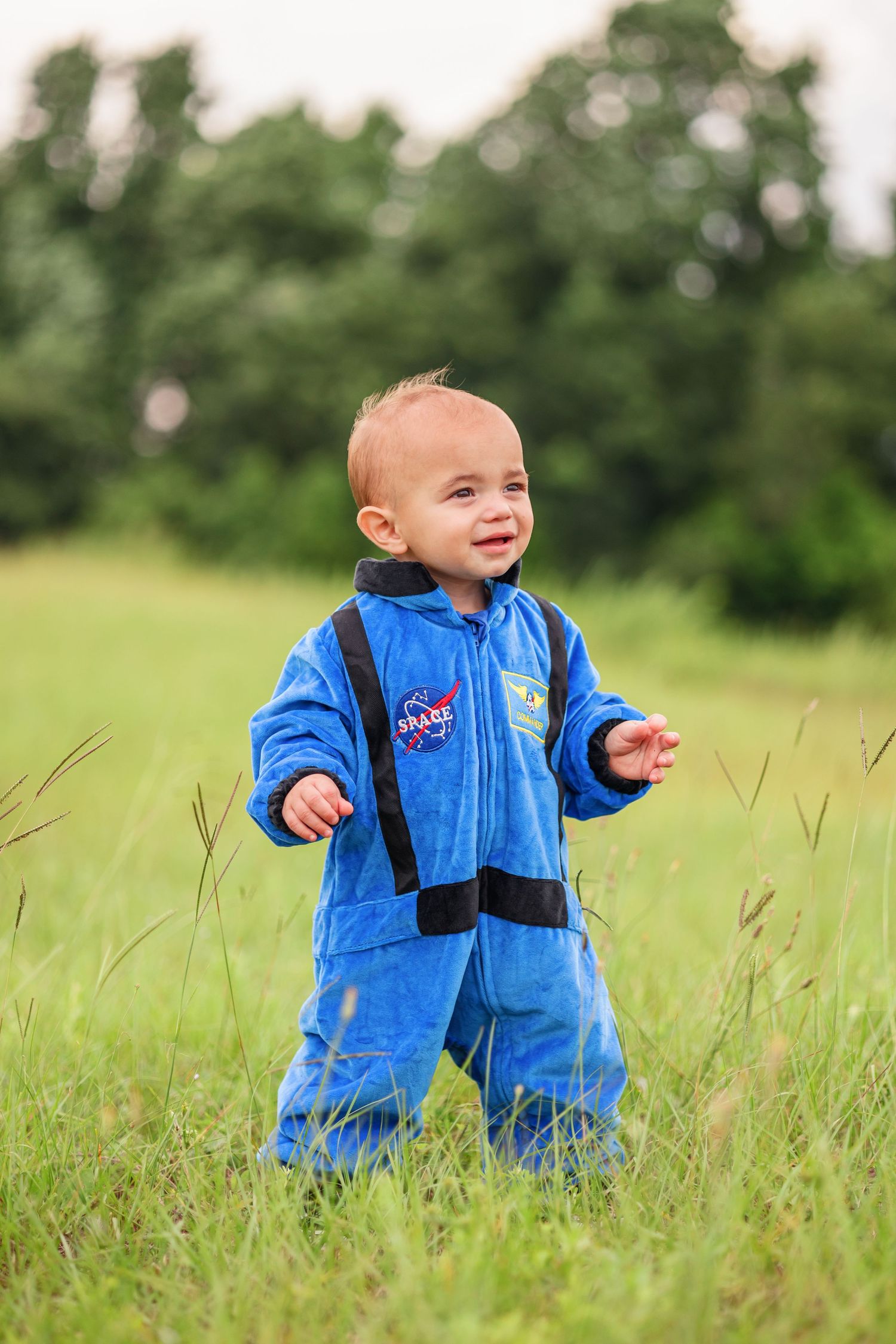 A toddler in a blue NASA spacesuit costume stands smiling in a grassy field.