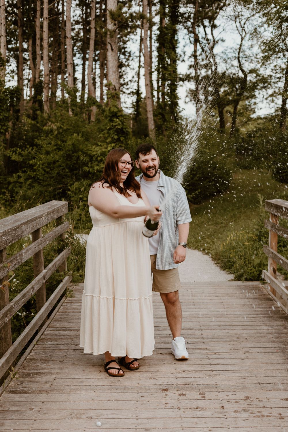 Happy couple popping champagne on a wooden bridge surrounded by lush green trees.