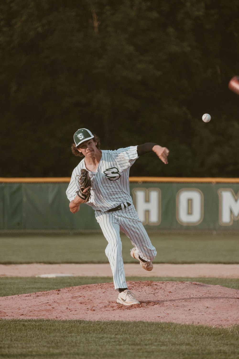 Baseball pitcher in green pinstripe uniform throwing a pitch on the mound at dusk.