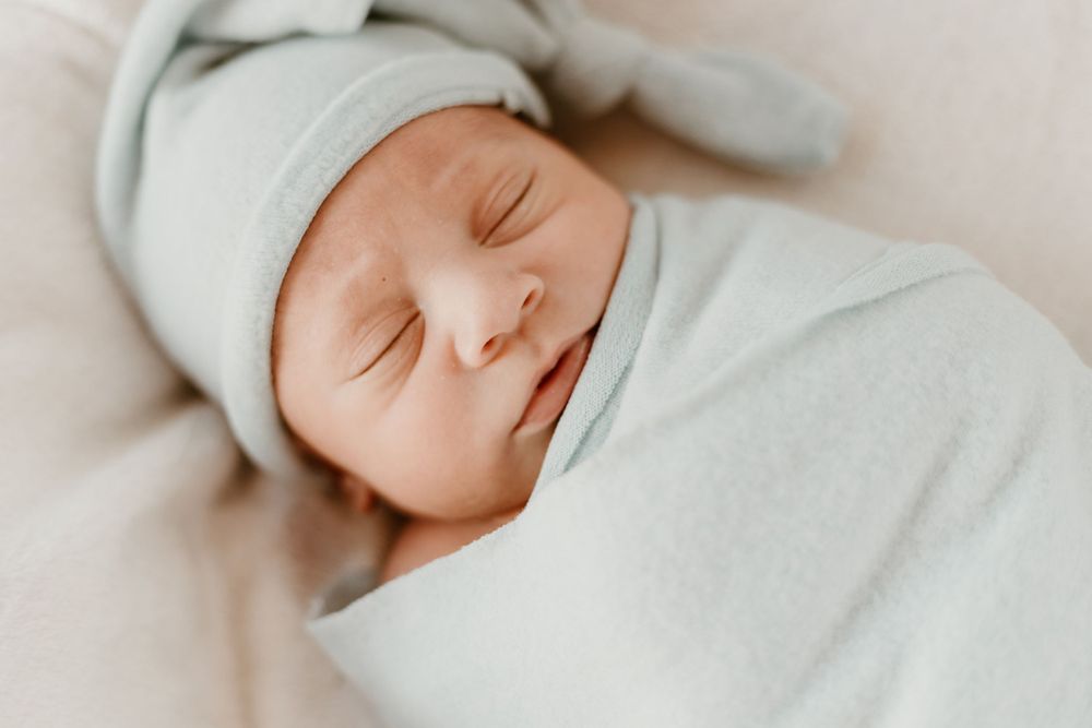 Peaceful newborn baby swaddled in white blanket wearing a knotted hat sleeping soundly.