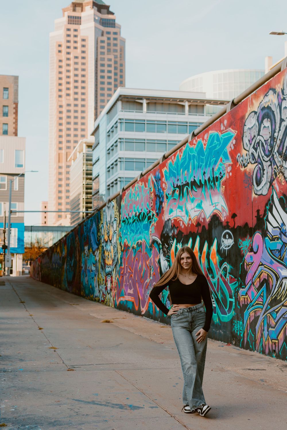 Young woman in black top and jeans poses confidently in front of a colorful urban graffiti wall.