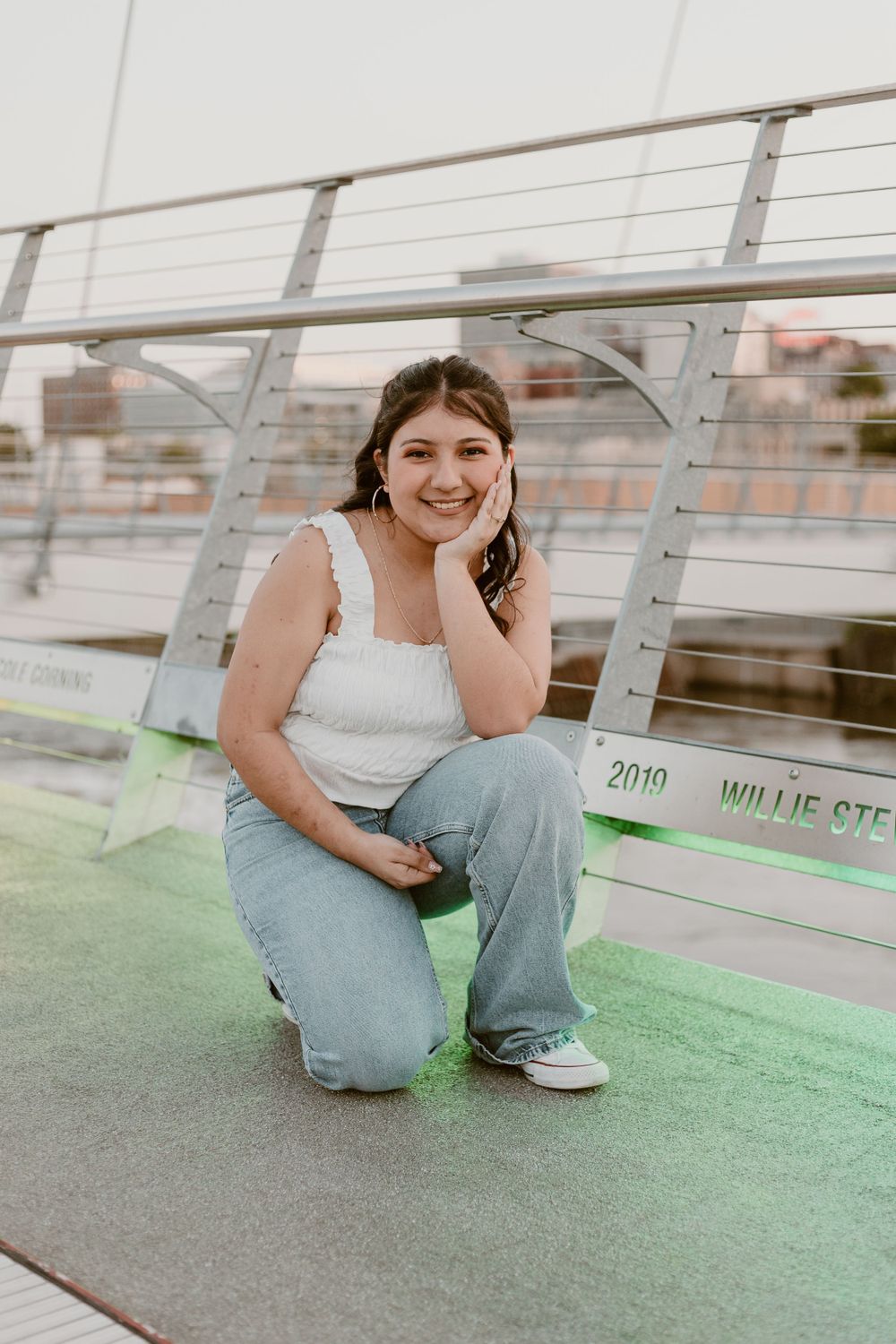Smiling young woman crouching on a green turf bridge in a white top and jeans.