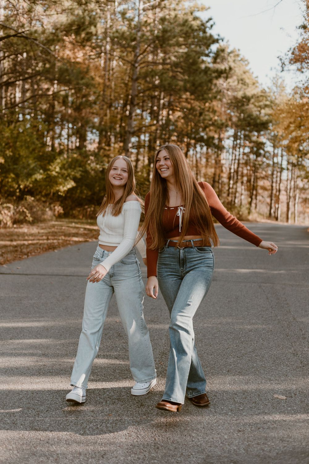 Two teenage girls laughing and posing together on a tree-lined road in autumn.