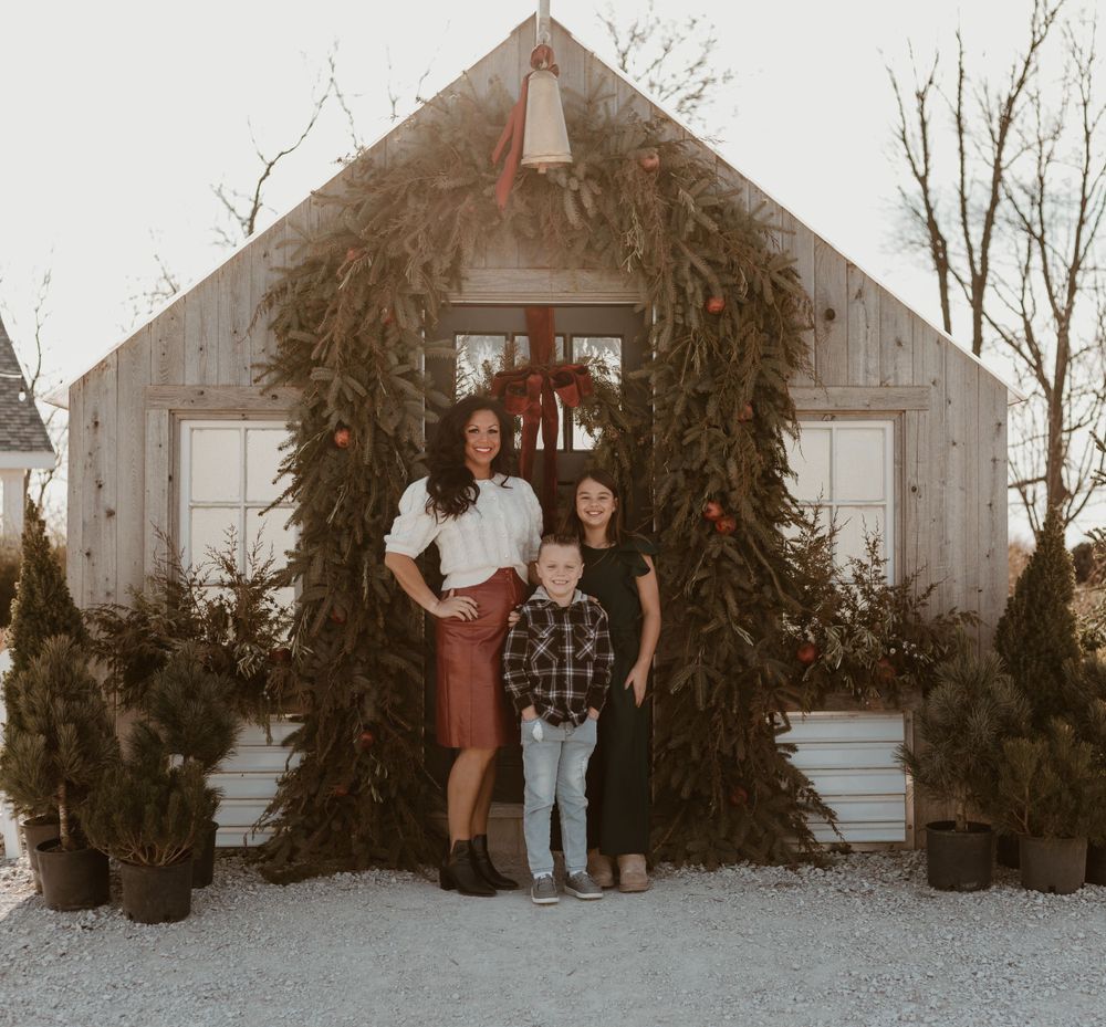 Mother and two children pose in front of a rustic barn decorated with Christmas wreaths and greenery.