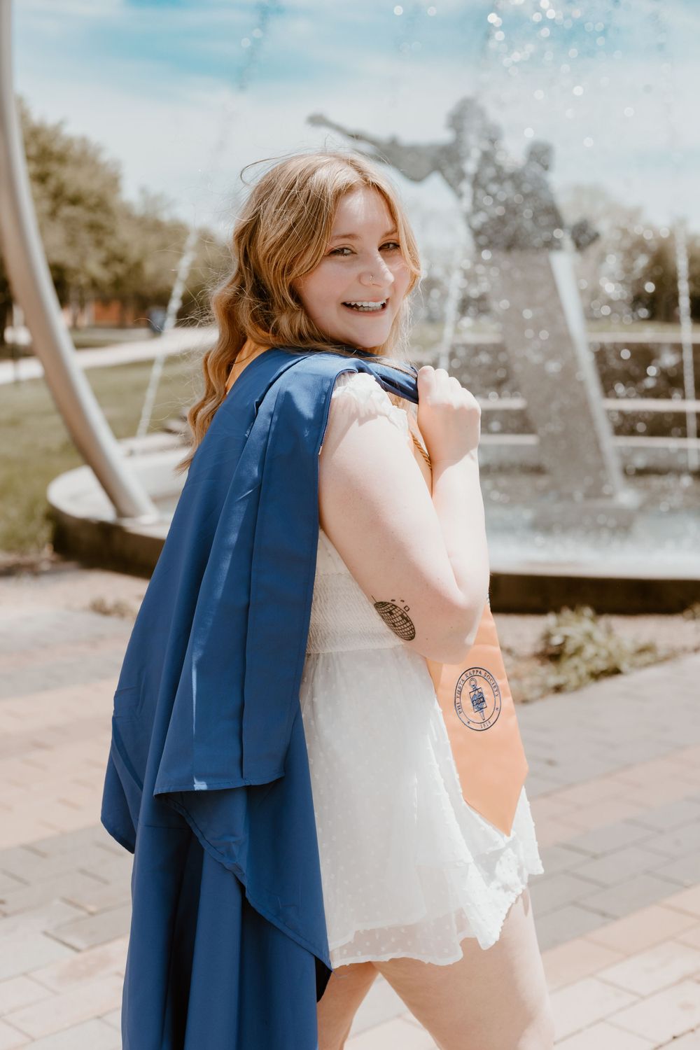 Smiling graduate in blue cap and gown holding sash near a fountain outdoors.