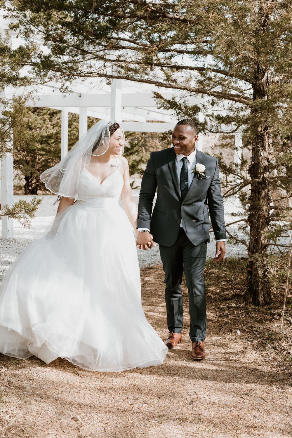 Bride and groom walking hand in hand near pine trees at an outdoor wedding venue.