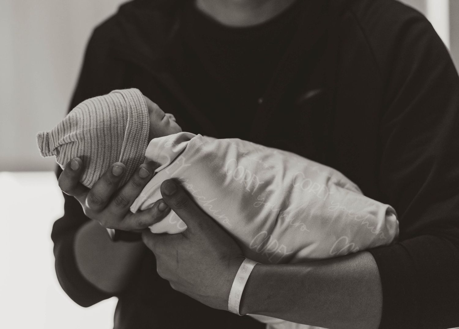 A black and white photograph shows a newborn baby swaddled in white cloth being cradled in someone's arms.