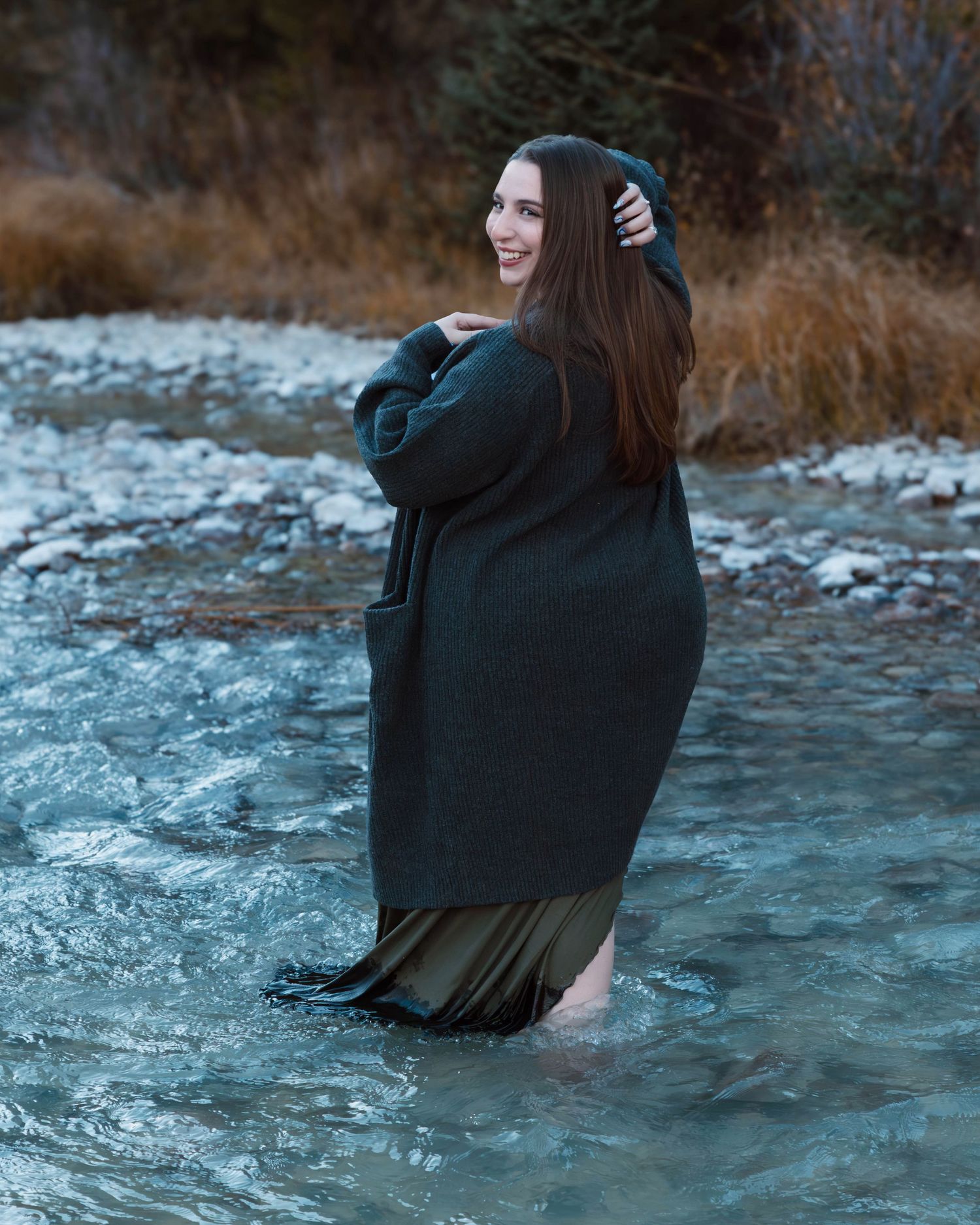 Artistic outdoor portrait in winter stream with flowing dark dress and cardigan.