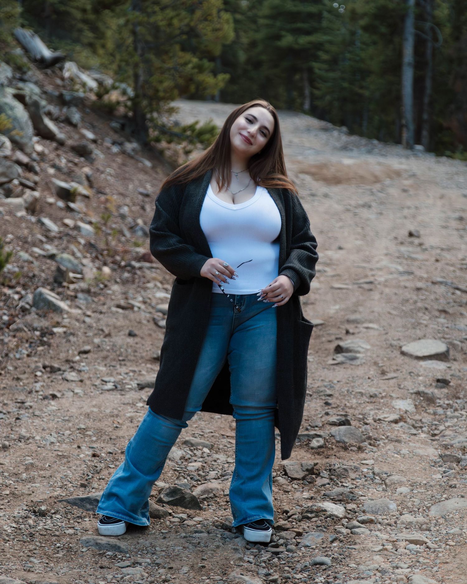 Someone poses on a mountain trail wearing a long black cardigan, white top and flared jeans against a forested backdrop.