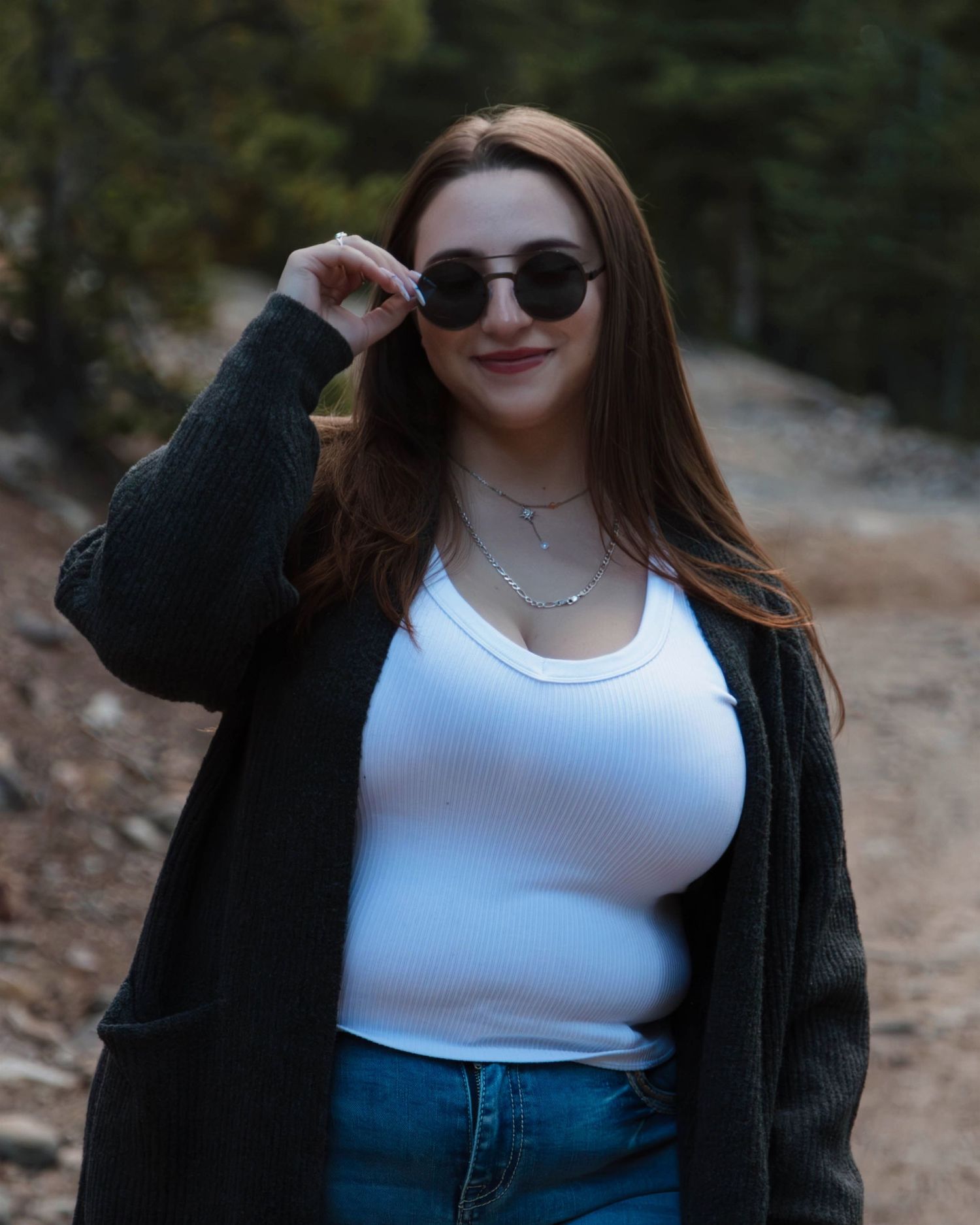 Person wearing sunglasses, white tank top and black cardigan poses outdoors on autumn trail with blurred forest background.
