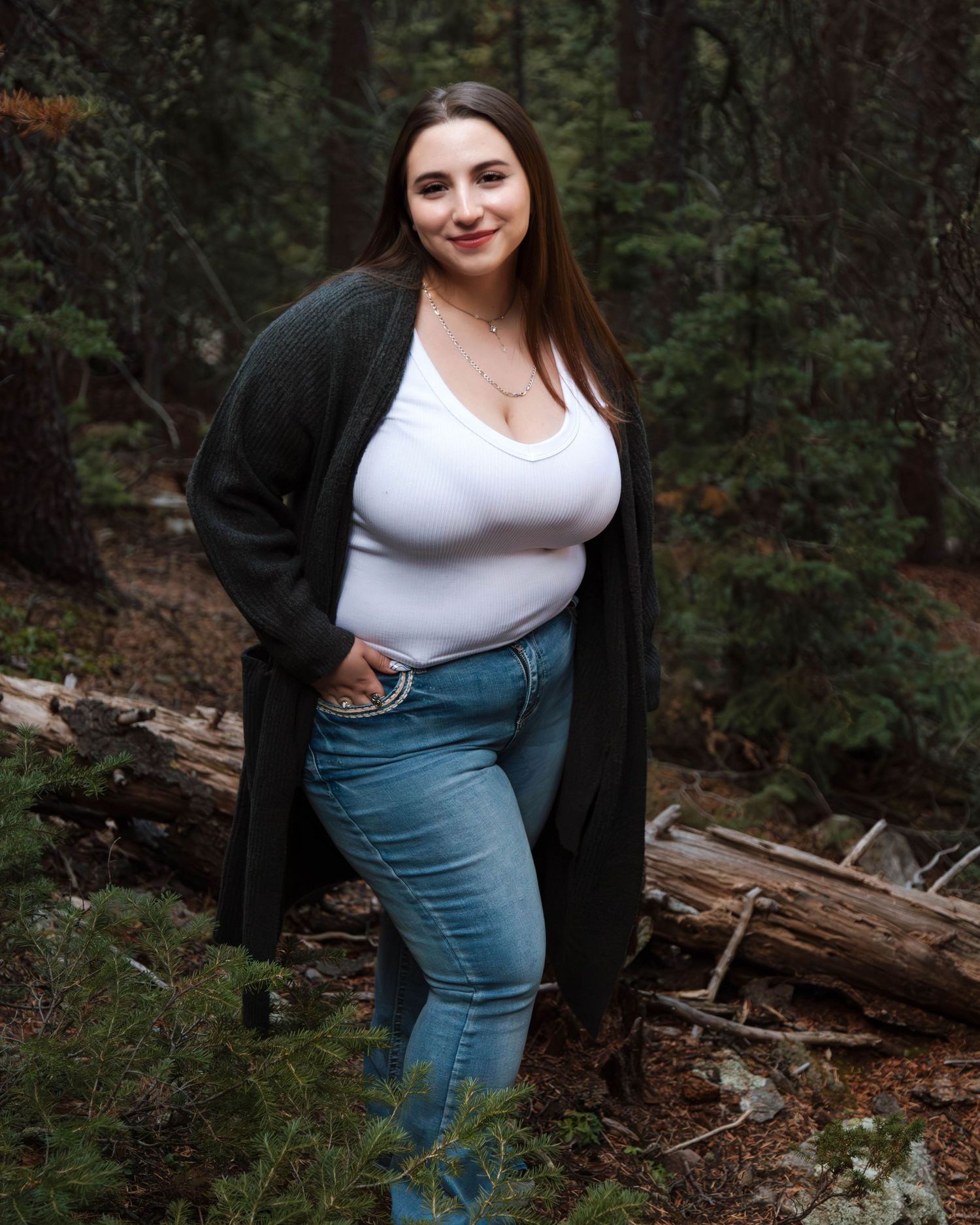 A woman in a black cardigan and white top poses in a wooded forest setting.