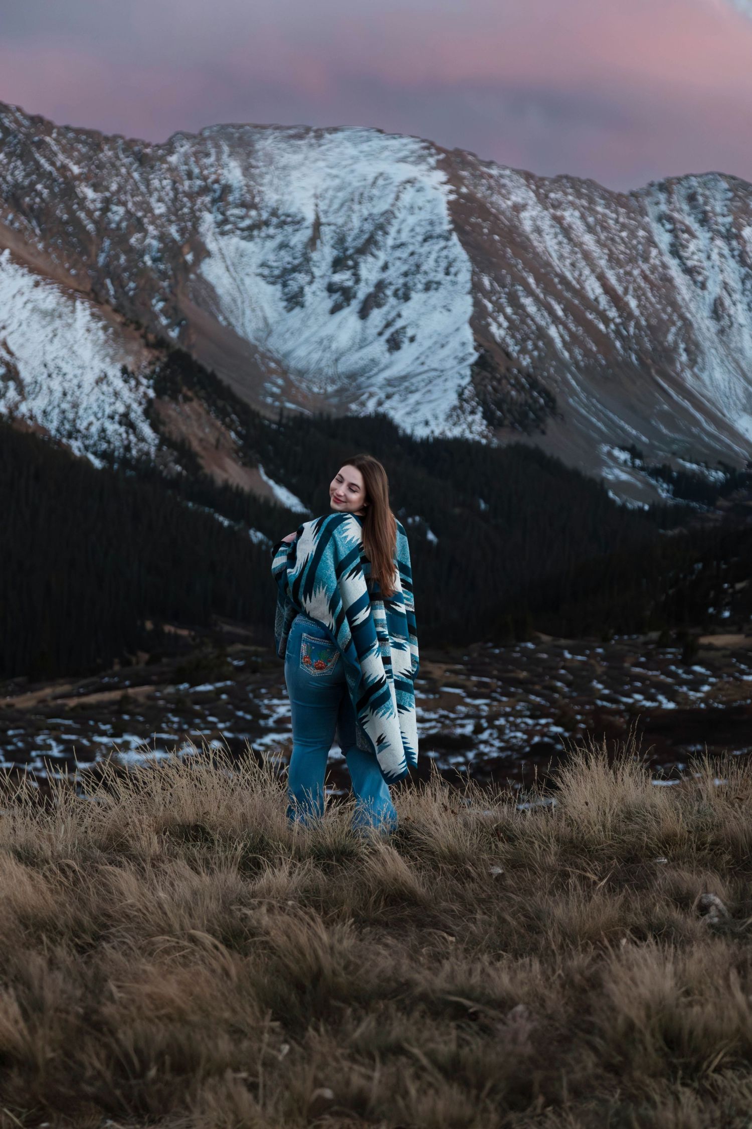 A person in a patterned jacket stands against a backdrop of snow-capped mountains during sunset.