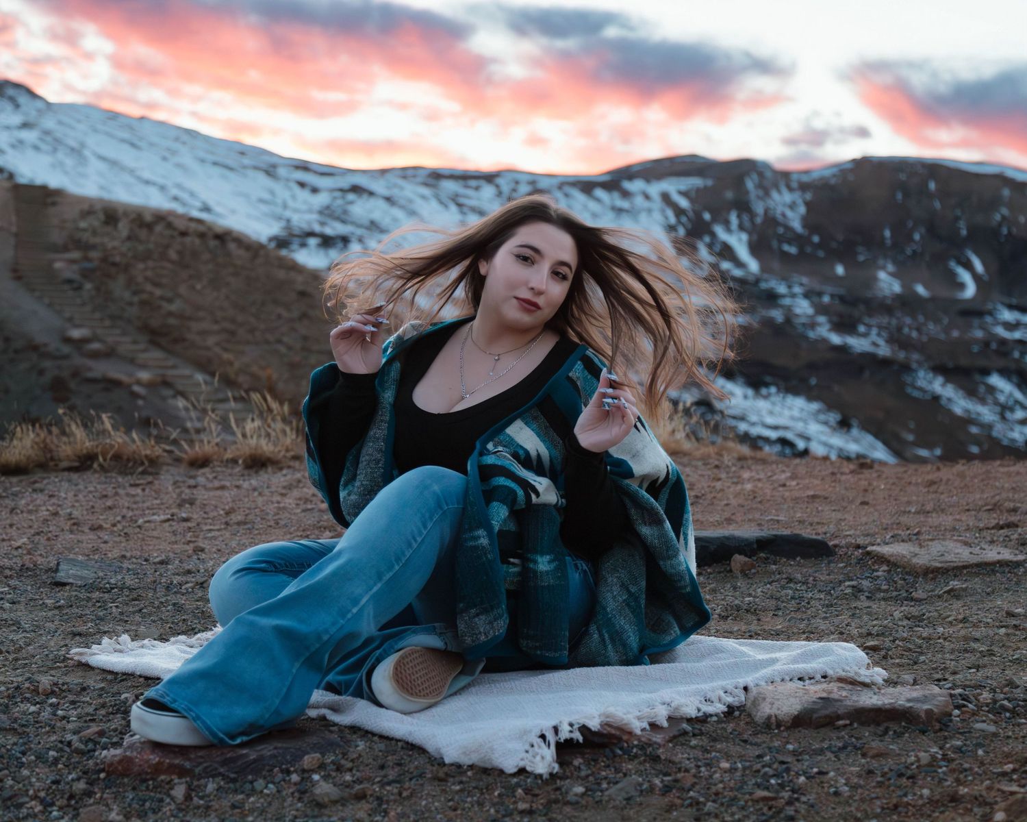 Person in denim sits on blanket against snowy mountain backdrop during colorful sunset.
