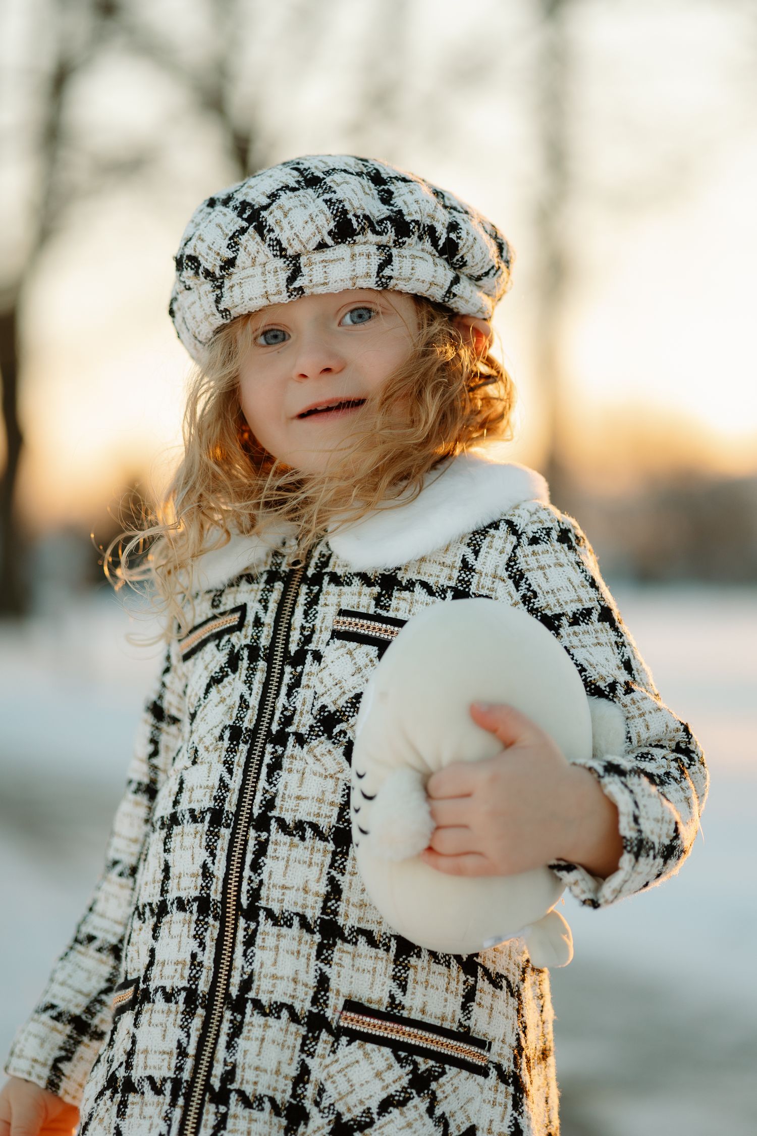 Young child in matching plaid coat and cap smiles while holding a white object in winter setting.