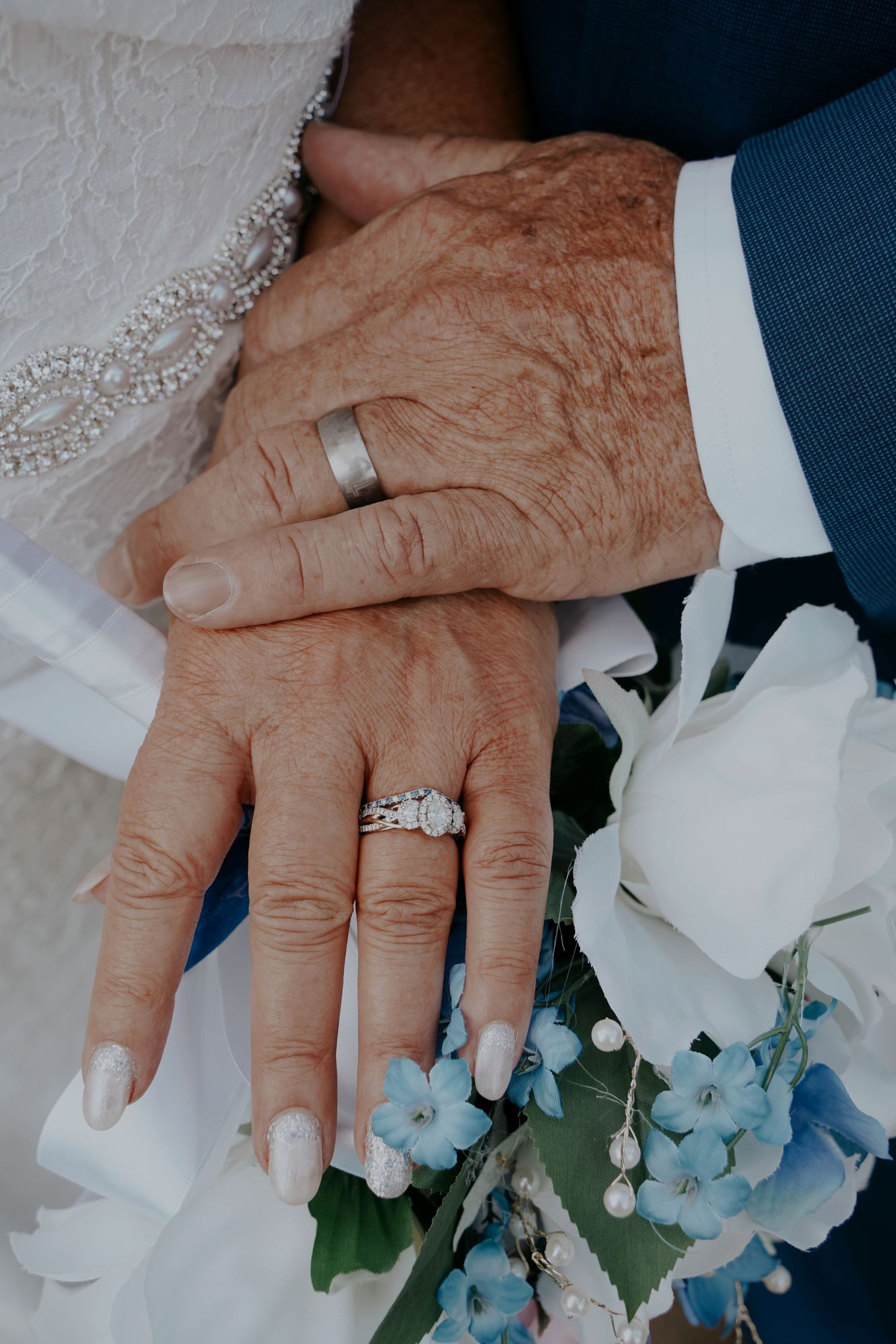 Close-up of elderly newlywed couple's hands showing wedding rings over a blue and white bouquet.