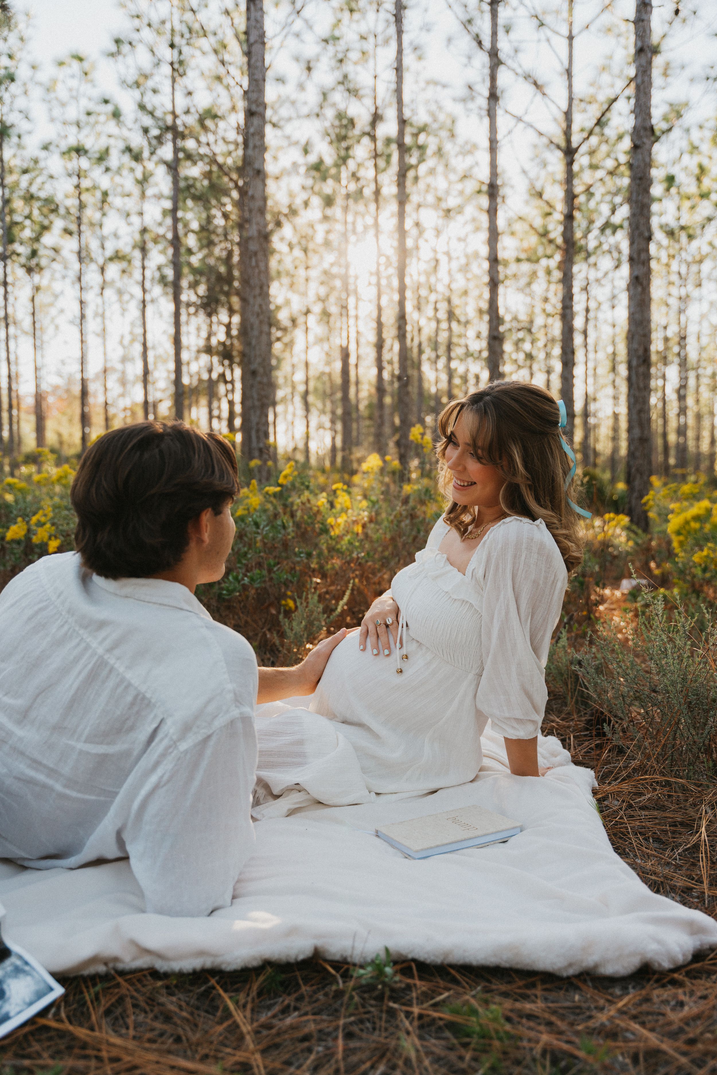 Romantic maternity photo session in a sunlit pine forest with couple sitting on white blanket among yellow wildflowers.