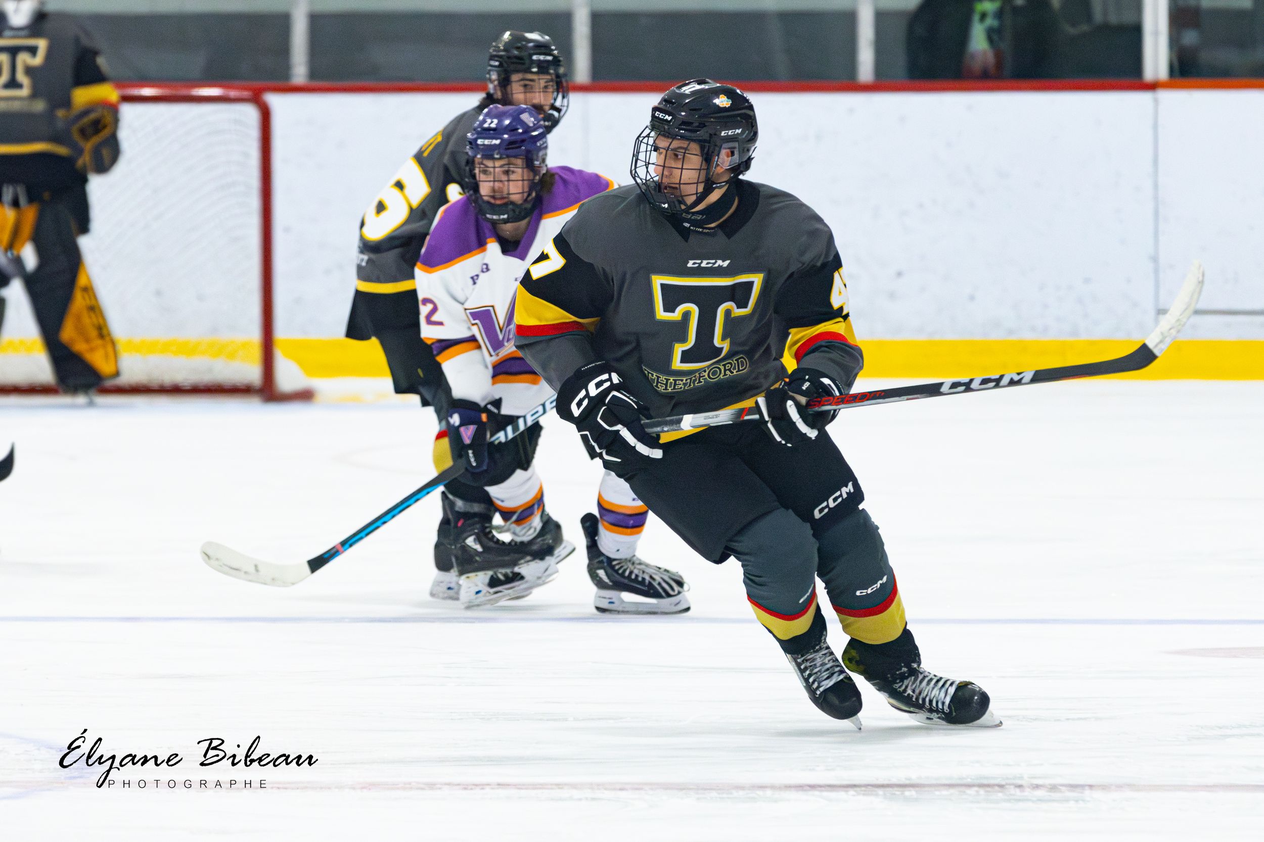 Hockey player in dark jersey skates with puck while opponent follows in purple.